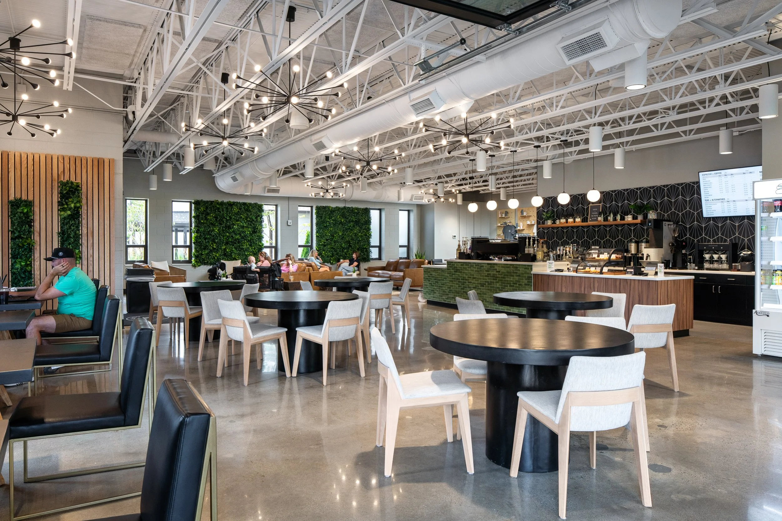 Wide view of cafe with black tables, wood chairs, greenery walls, and modern pendant lights.