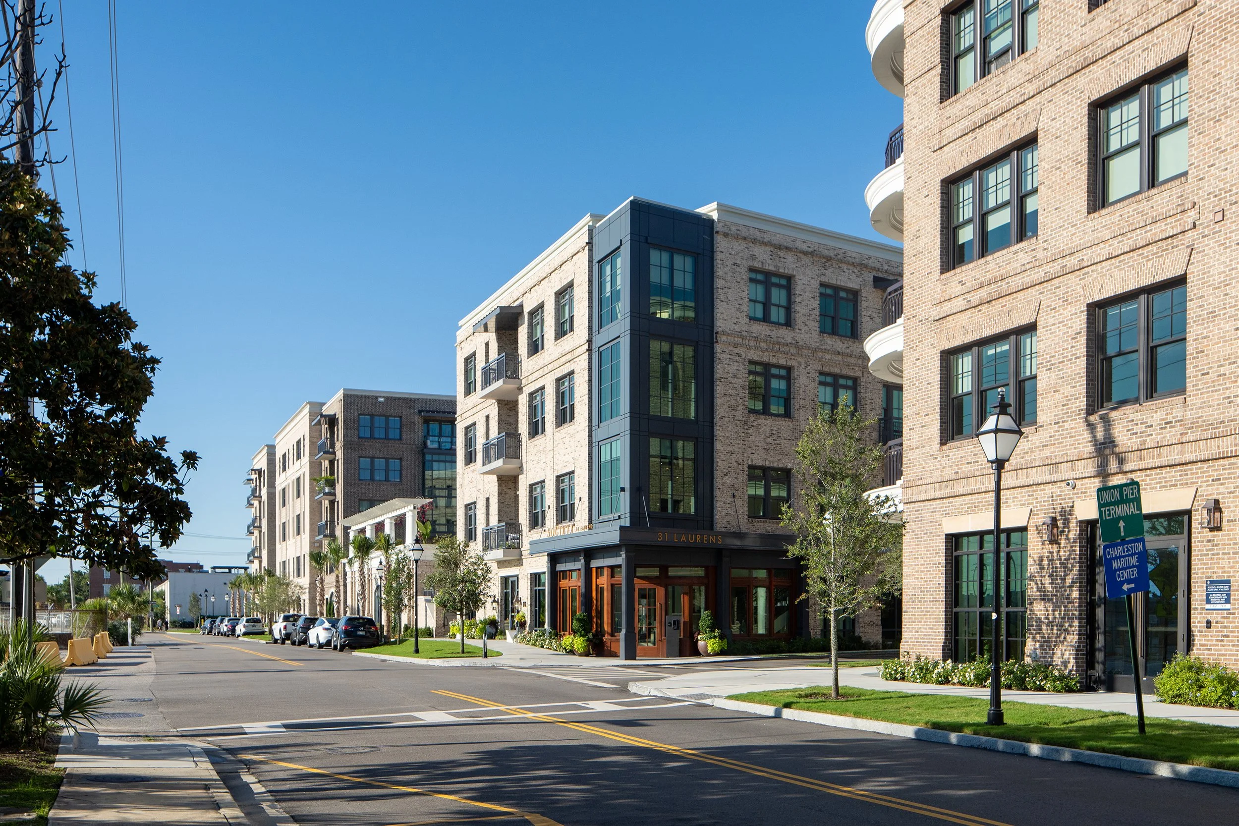 Street view down Laurens Street with the 31 Laurens entrance featuring wood-framed glass doors, the Society at Laurens building beyond, and a green Union Pier Terminal sign directing toward the Charleston Maritime Center.