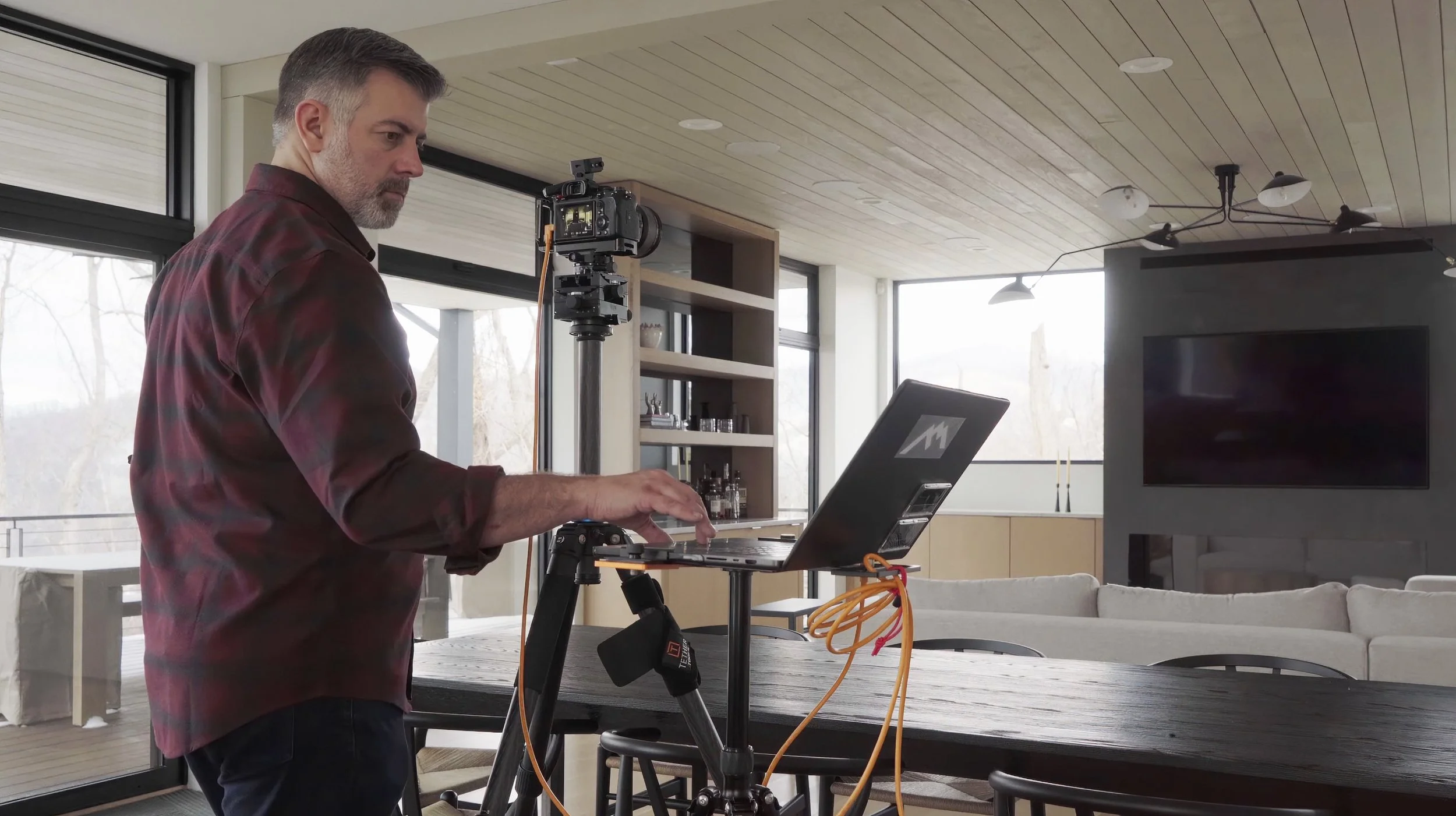 A man working on a laptop connected to a camera mounted on a tripod in a modern living room with large windows, wooden ceiling, and a television.