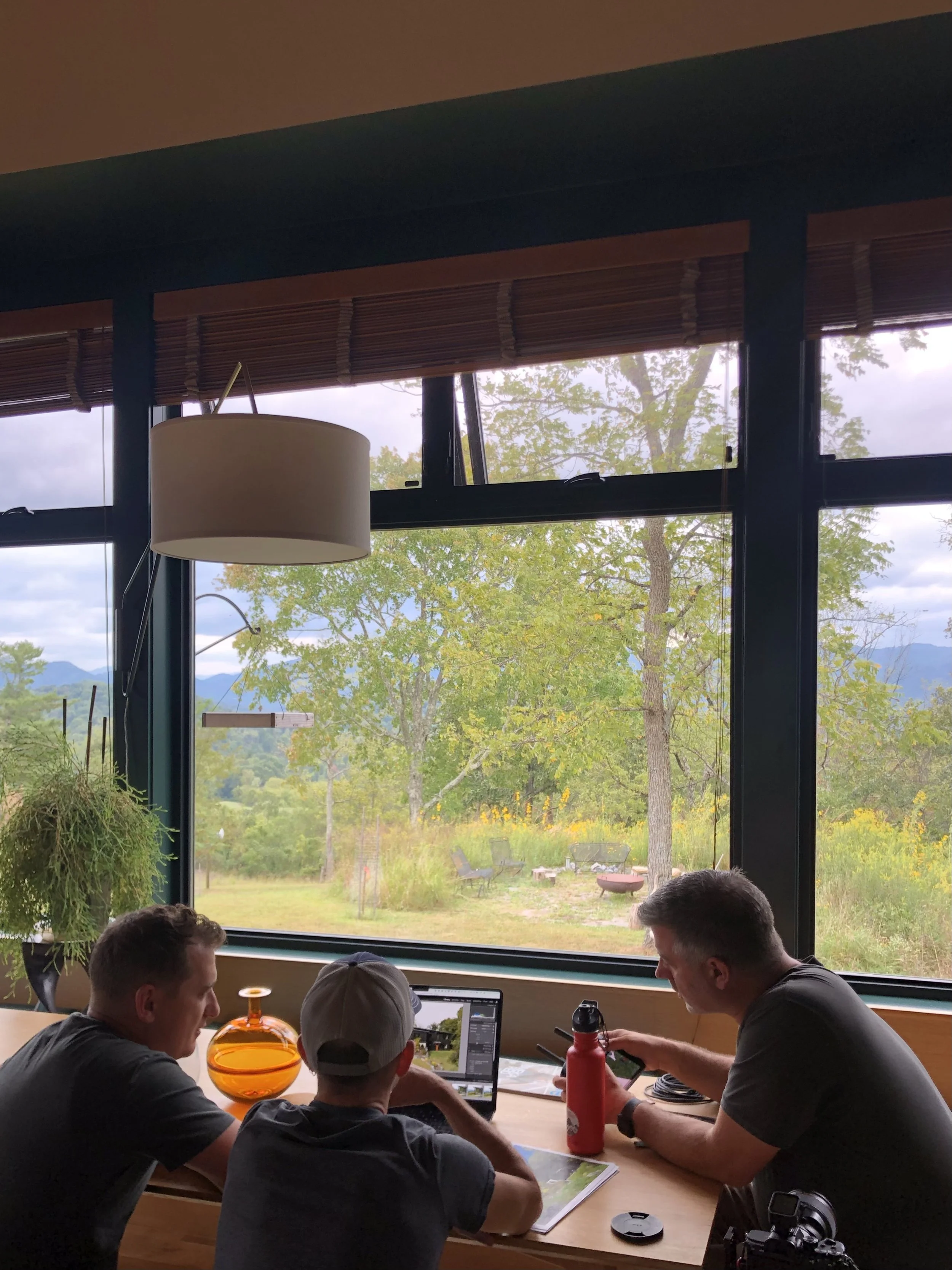 Group of four people sitting at a wooden table in a room with large windows, viewing a landscape with trees and mountains outside. They have laptops, drinks, and a camera on the table.