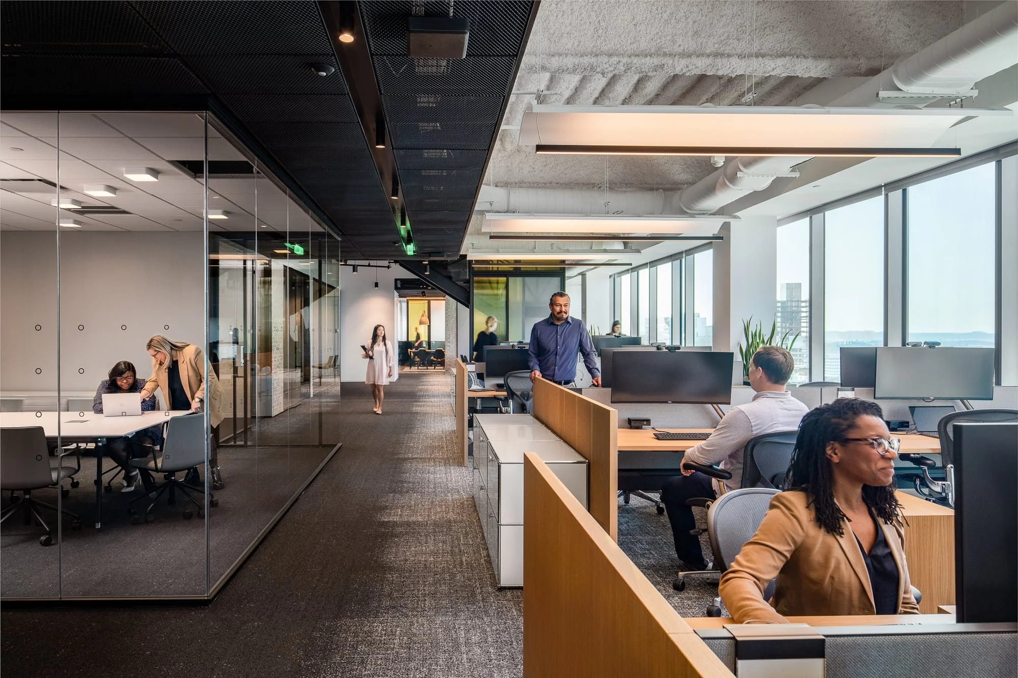 Alliance Bernstein Nashville open office floor with employees working at wood-paneled workstations, glass-walled meeting rooms, exposed ceiling, and city views beyond
