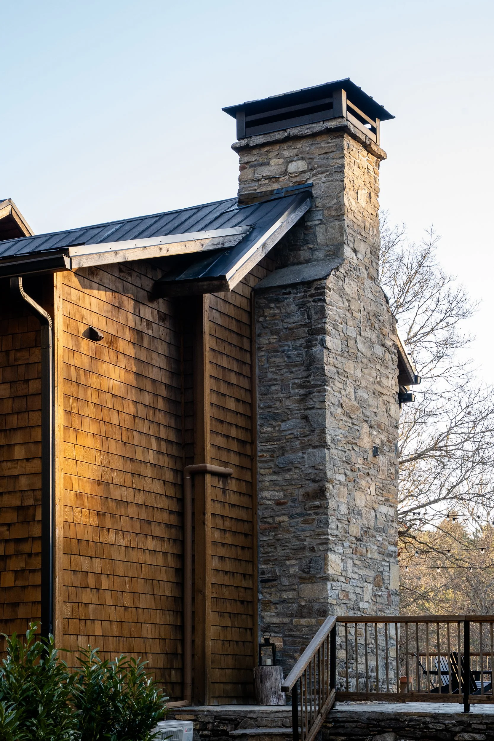 Close view of a tall stone chimney beside cedar shingles and a dark metal roof.