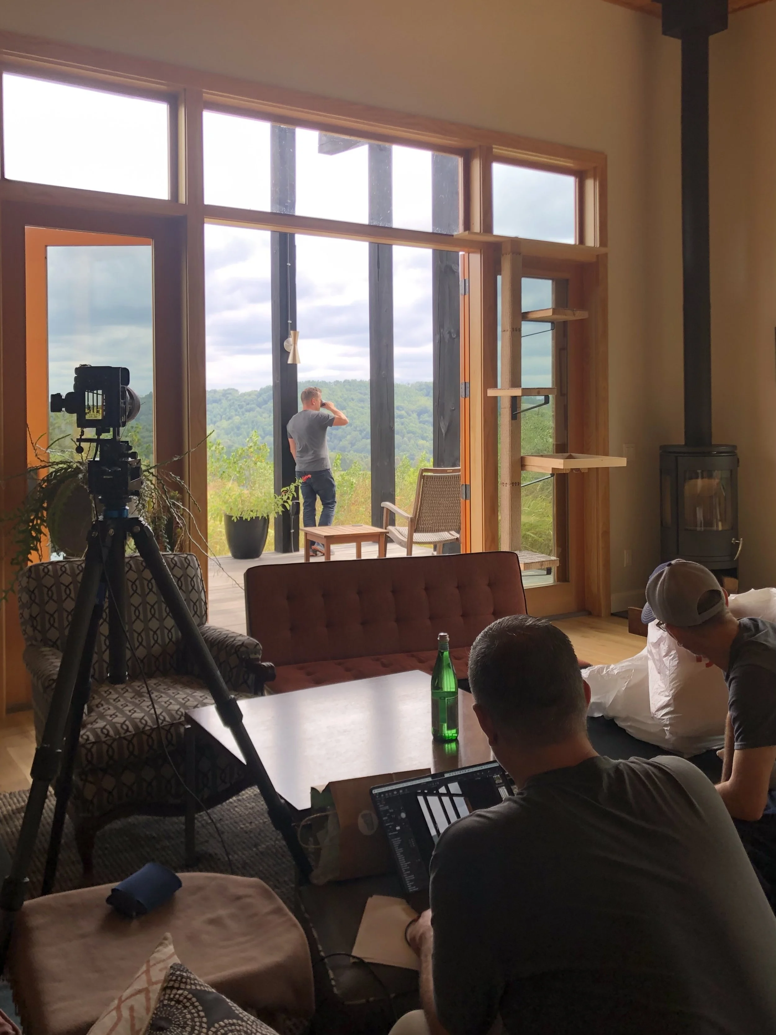 A group of people inside a room with large windows showing a green outdoor landscape, some individuals working with cameras and laptops, and others sitting or standing near a wooden table and a cozy stove.