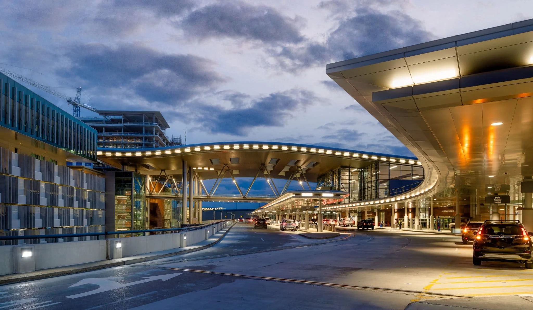 Twilight view of an airport departures roadway beneath a sweeping illuminated canopy with terminal glass, traffic, and drop-off lanes.