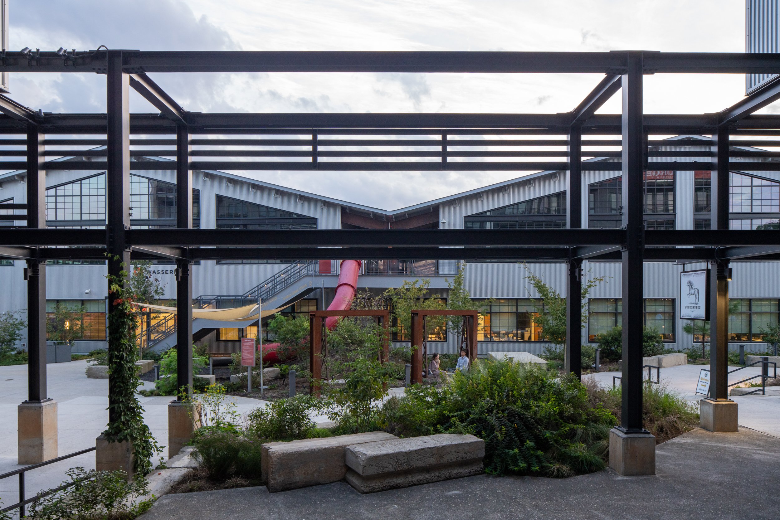 View through a black steel pergola toward the campus interior, with a red tube slide spanning between levels, the Ponysaurus brewery storefront, and visitors gathered beneath weathering steel arches.