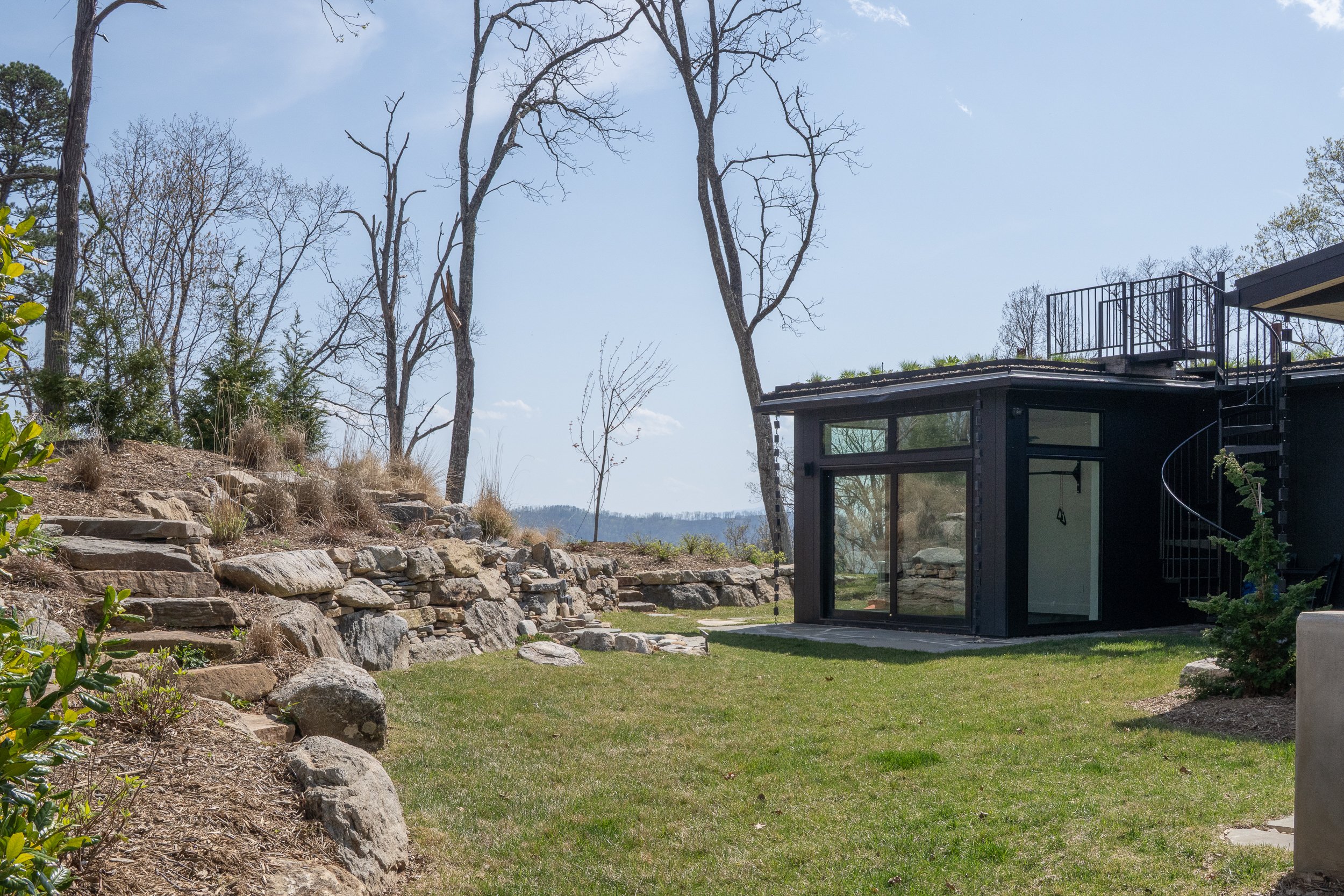 Lower porch and glass-walled room beside stone terraces, lawn, and mountain views.