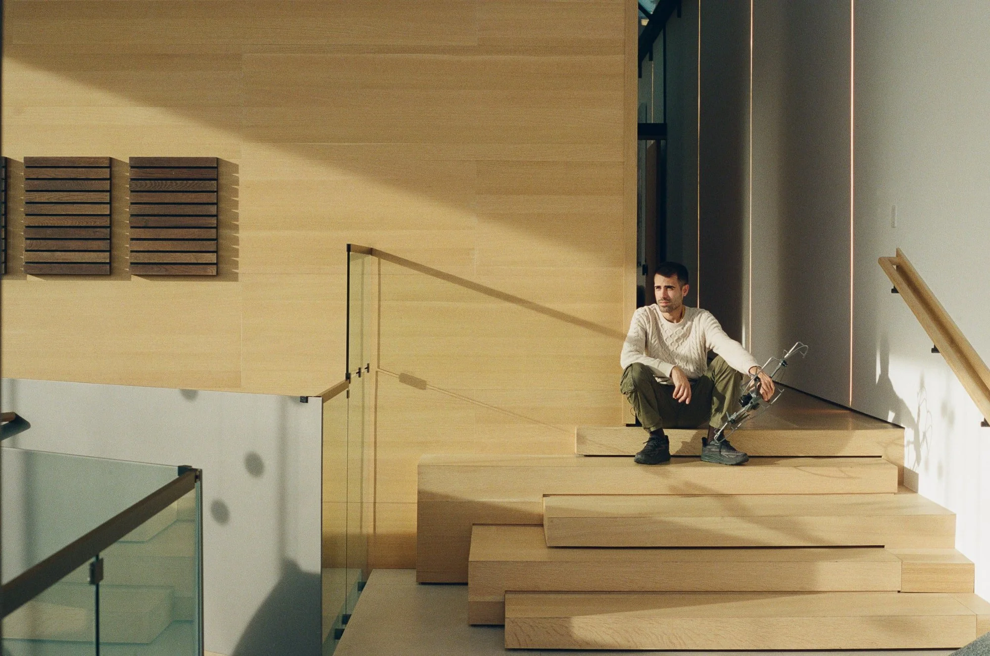 Person seated on a stepped wood stair beside a glass railing, warm wood walls, and late-day sunlight.