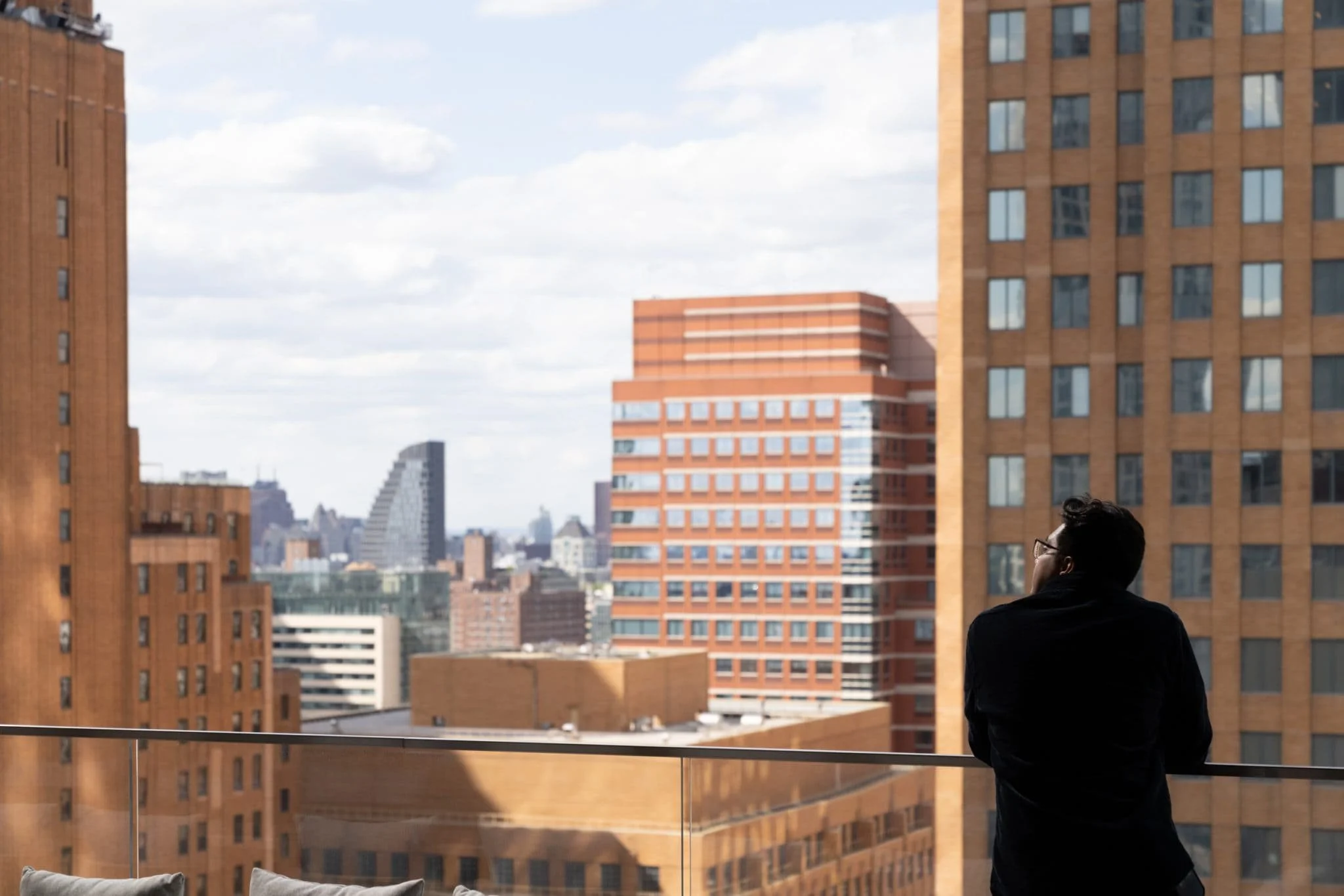 Person leaning on a glass railing of a high-rise terrace with brick buildings and a city skyline in the distance.
