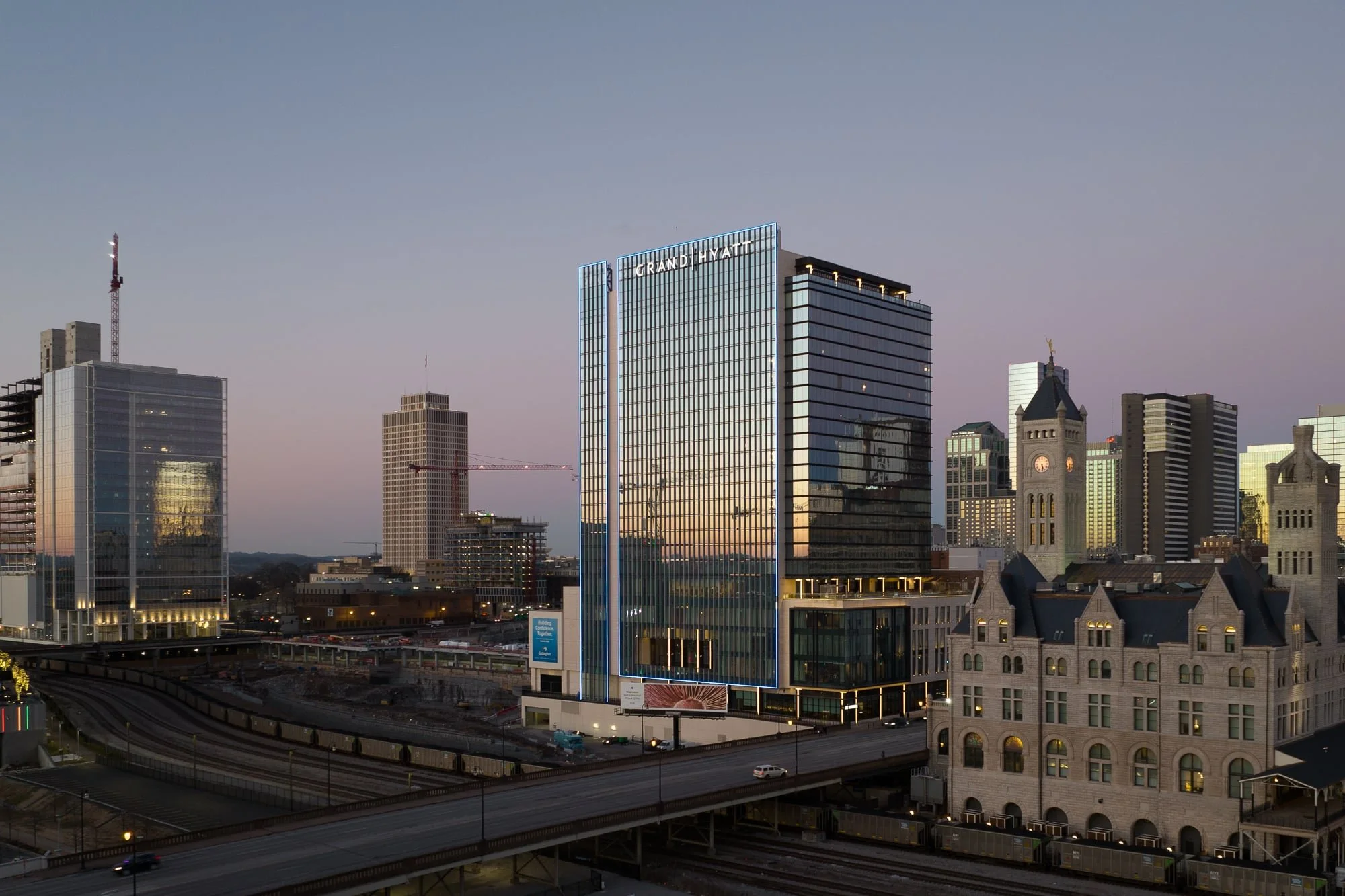 Aerial drone view of the Grand Hyatt at Nashville Yards at twilight with a tall reflective glass tower, illuminated signage, surrounding downtown architecture including Union Station, and rail tracks below