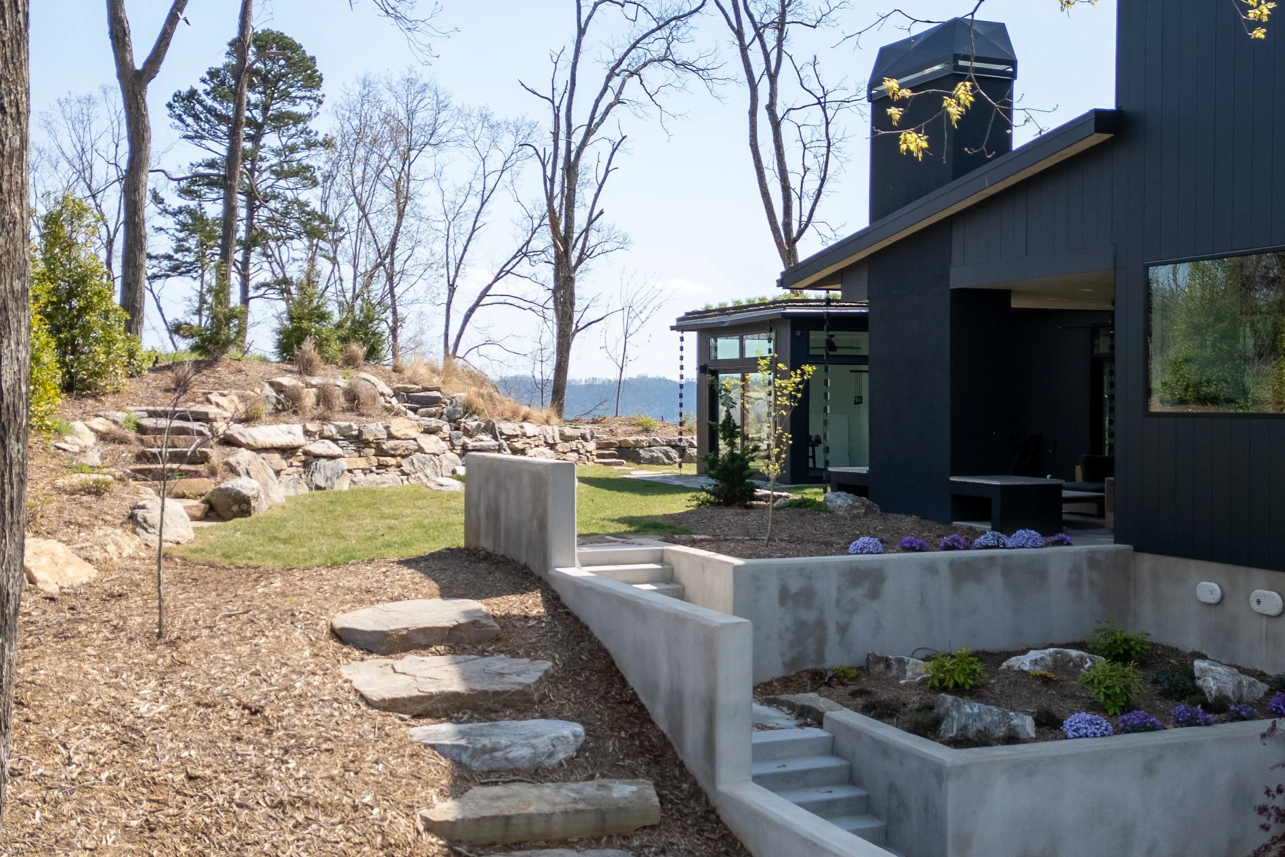 Courtyard landscape with stone steps, concrete walls, planting beds, and black exterior walls.