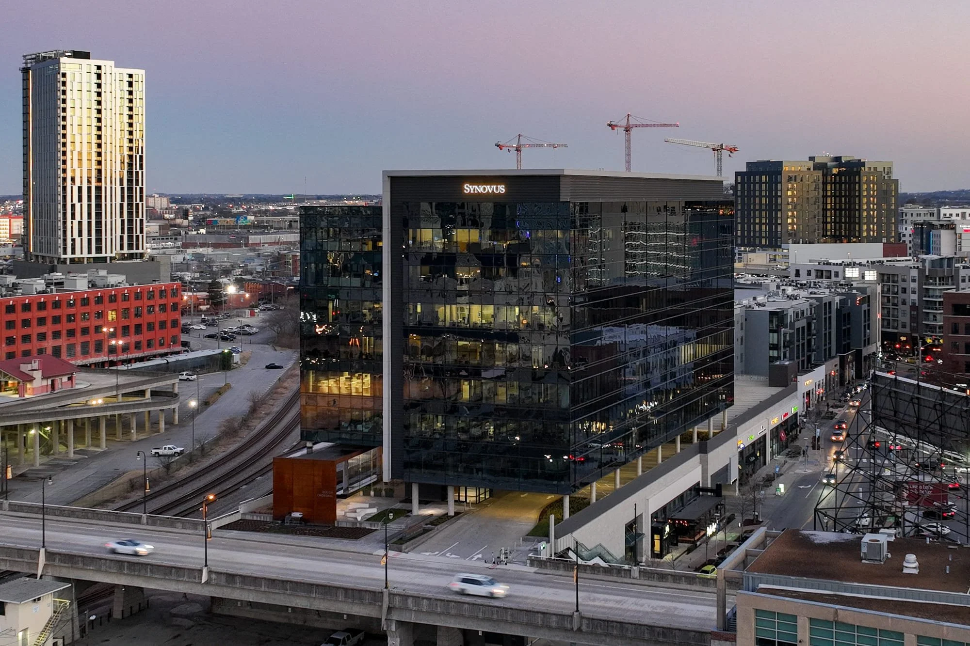 Aerial drone view of Gulch Crossing at dusk with the Synovus signage at the top, reflective glass facade, surrounding Gulch streetscape, freight rail line, and a residential tower in the background