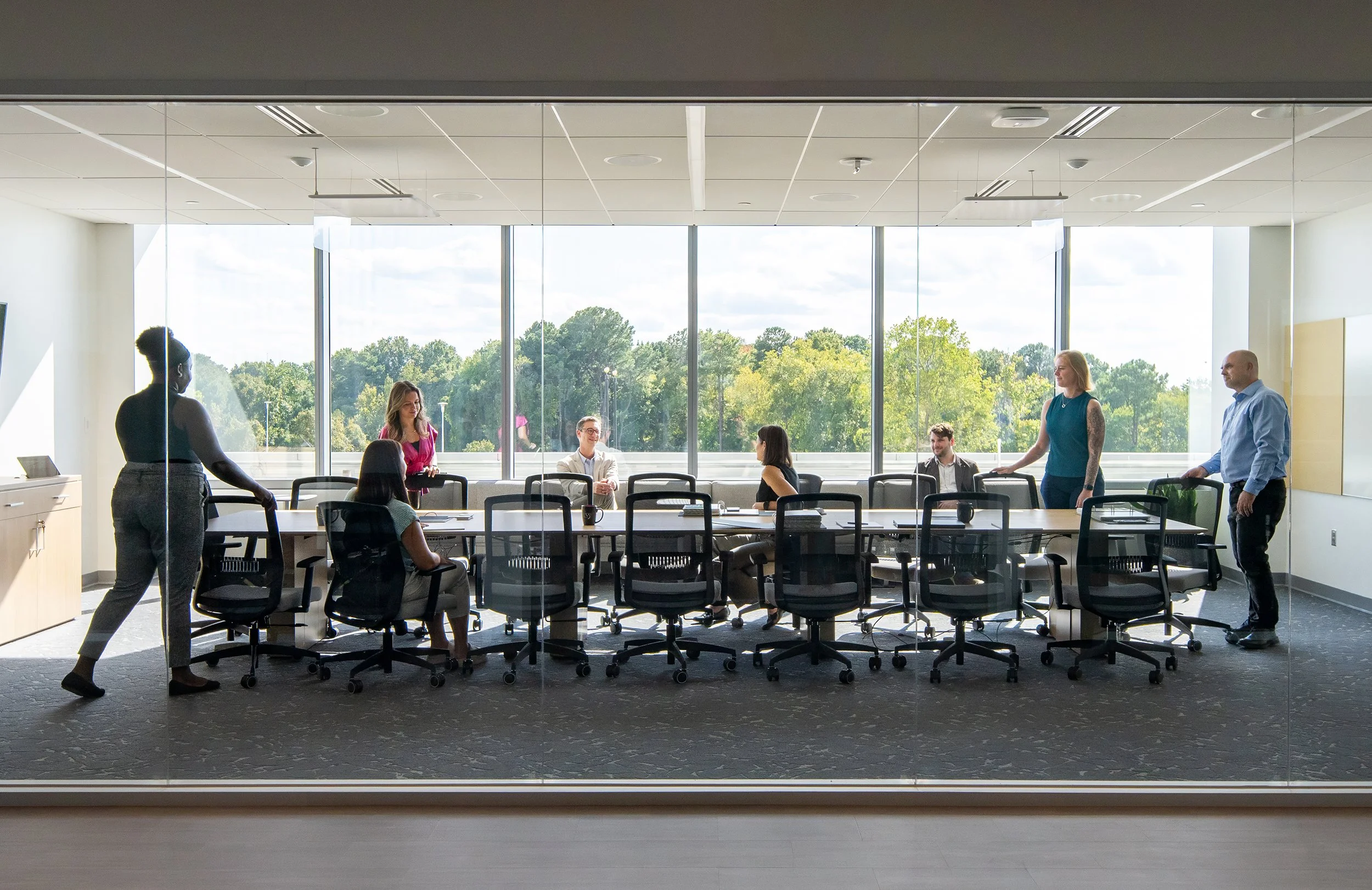 Conference room behind a full-height glass partition, with staff gathered around a long table backlit by floor-to-ceiling windows looking out to mature trees. 