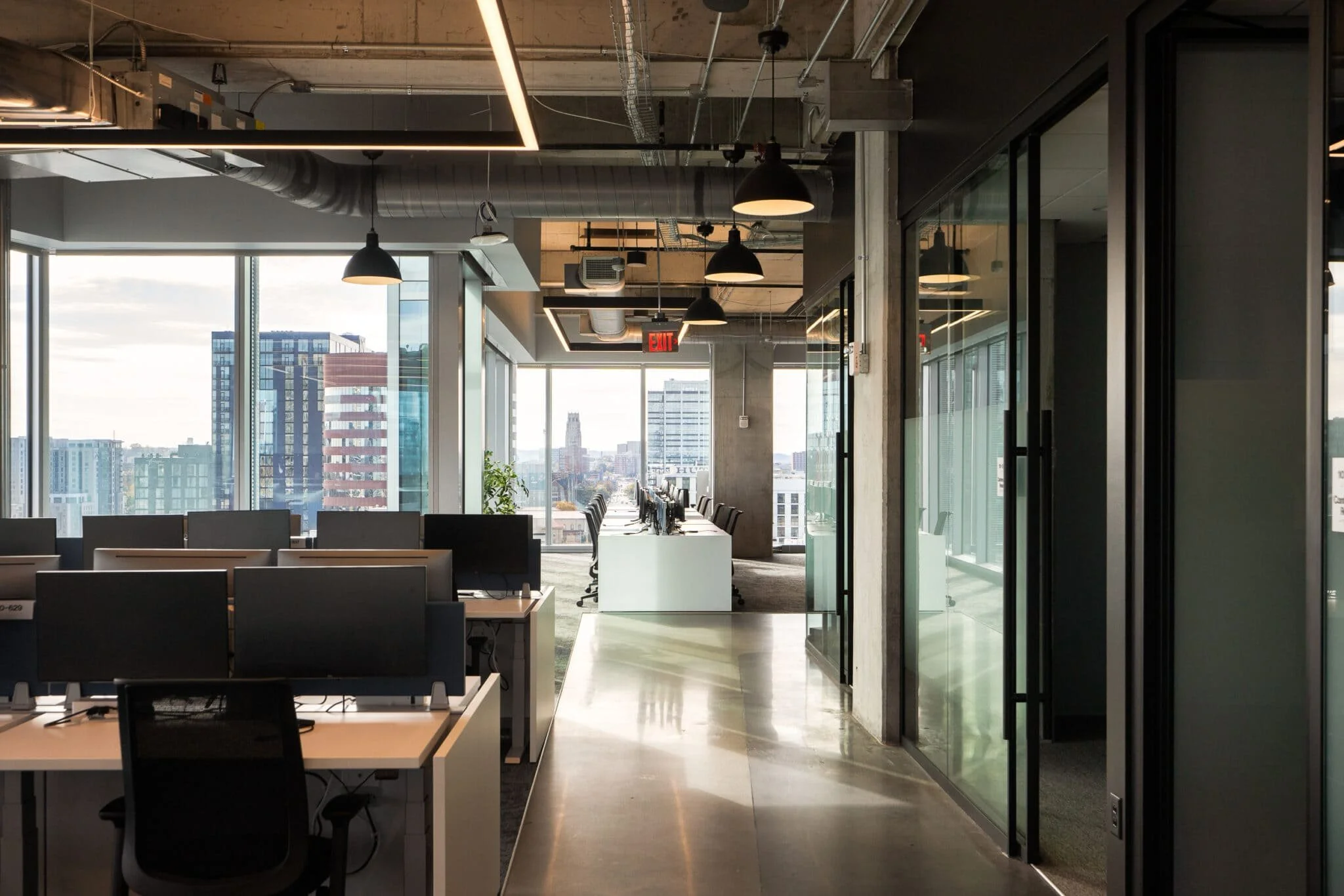 Open office with desk rows, glass meeting rooms, exposed ductwork, and late-day light from large windows.