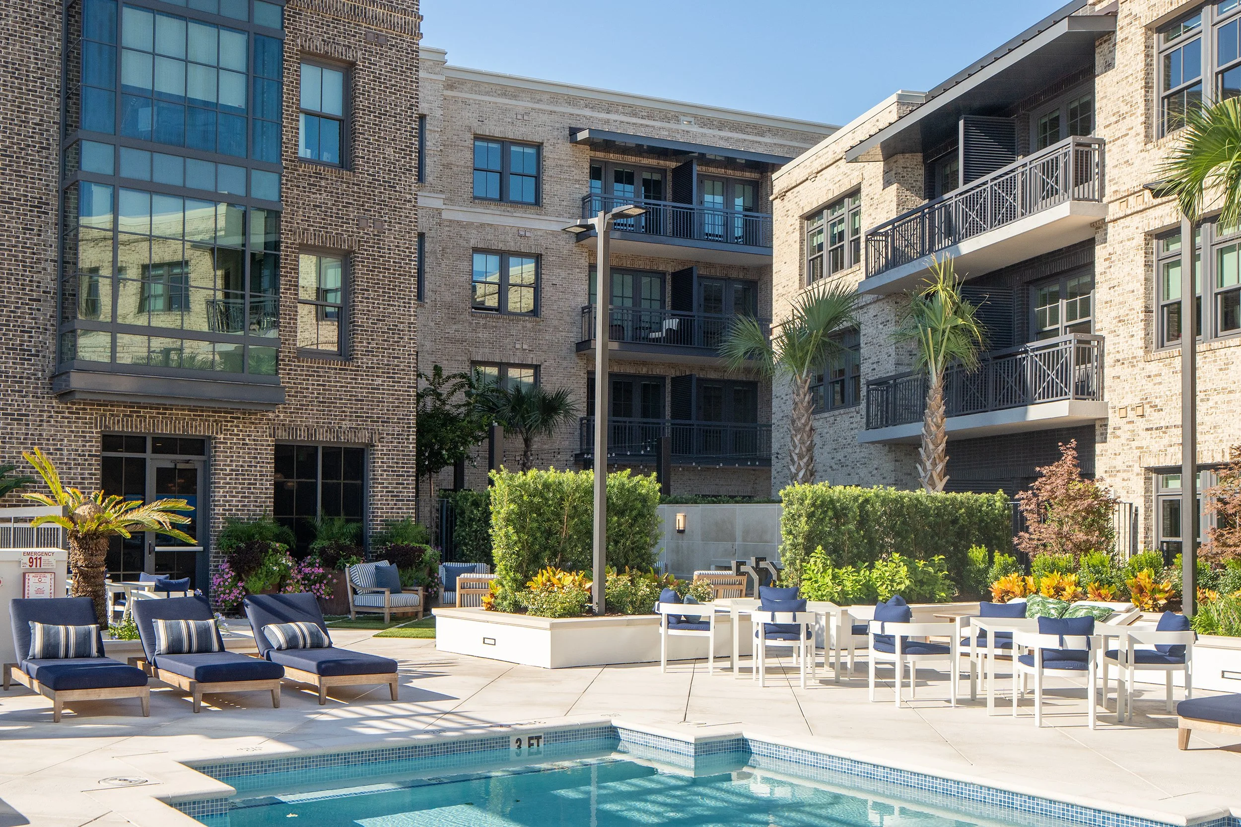 Pool courtyard with navy chaise loungers, white outdoor dining furniture, palm trees and clipped hedges, surrounded on three sides by brick residential buildings with stacked balconies.