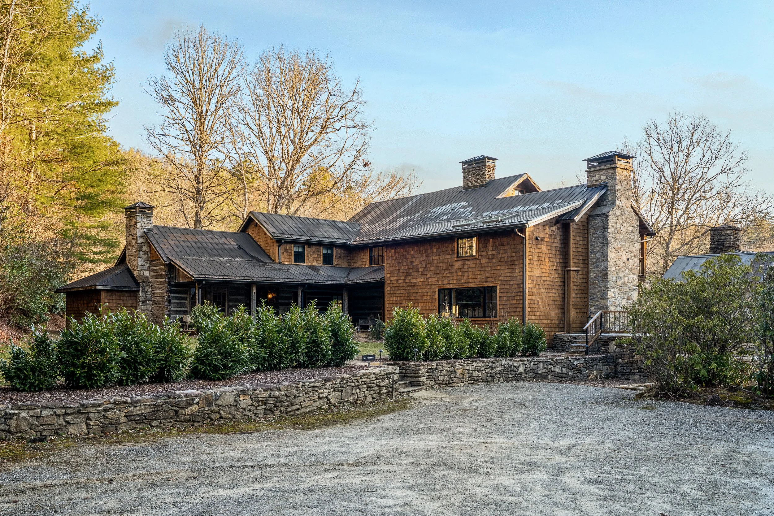 Gravel drive and stone walls leading to a shingle-sided mountain lodge with metal roofs and stone chimneys.