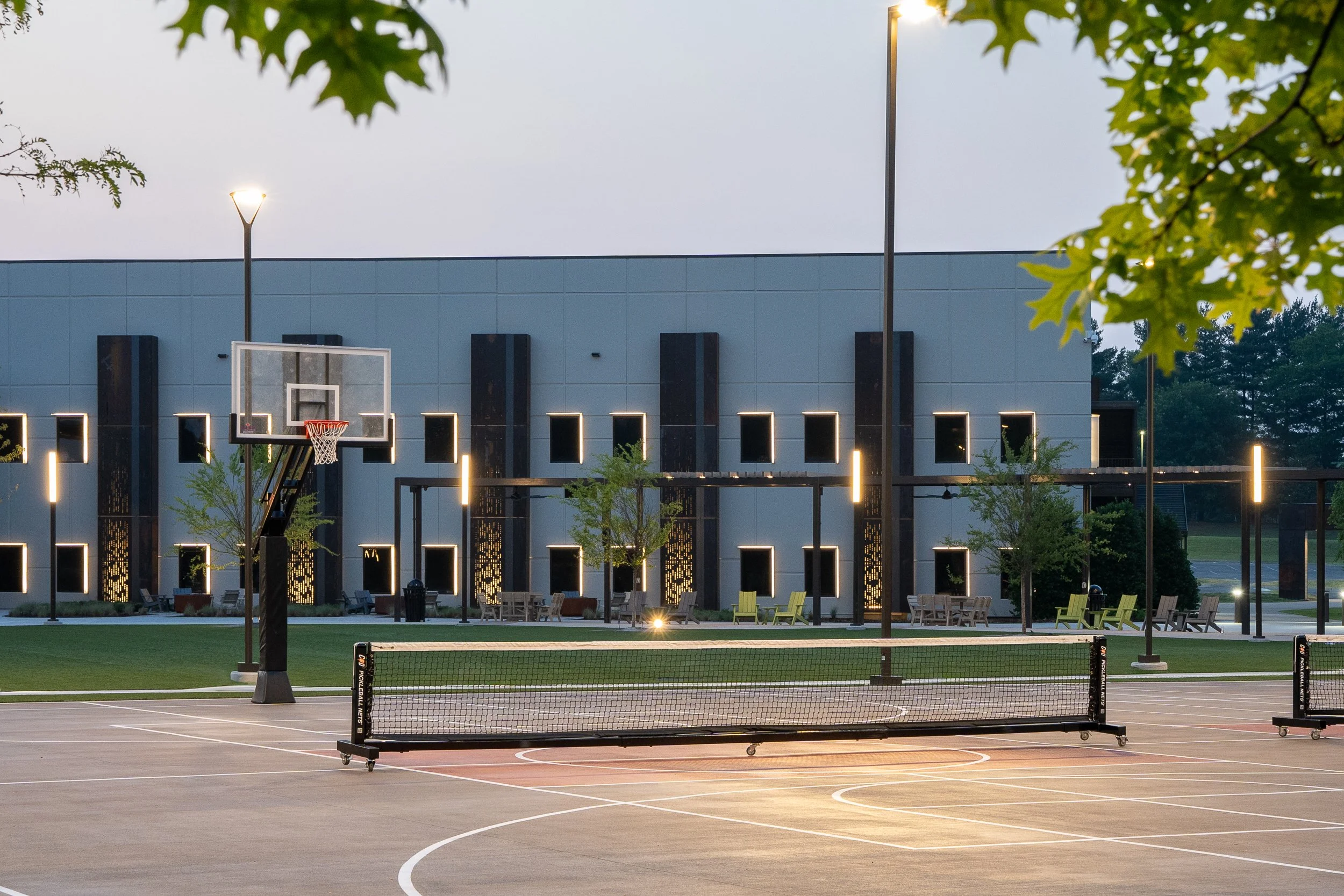 Outdoor basketball court at dusk with illuminated backboard, benches, and modern building facade.