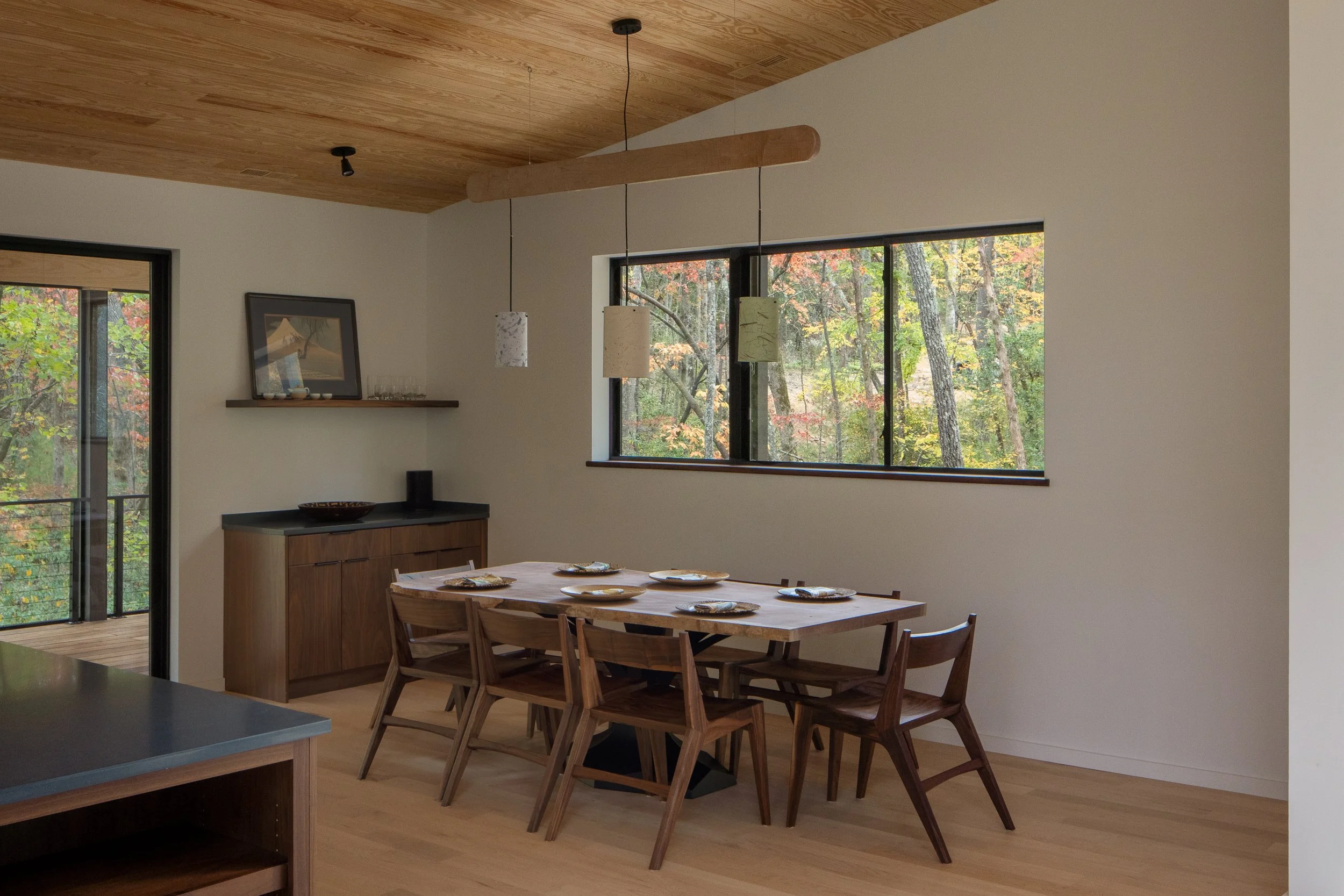 Dining area with a wood table, pendant light, and wide window looking into the forest.