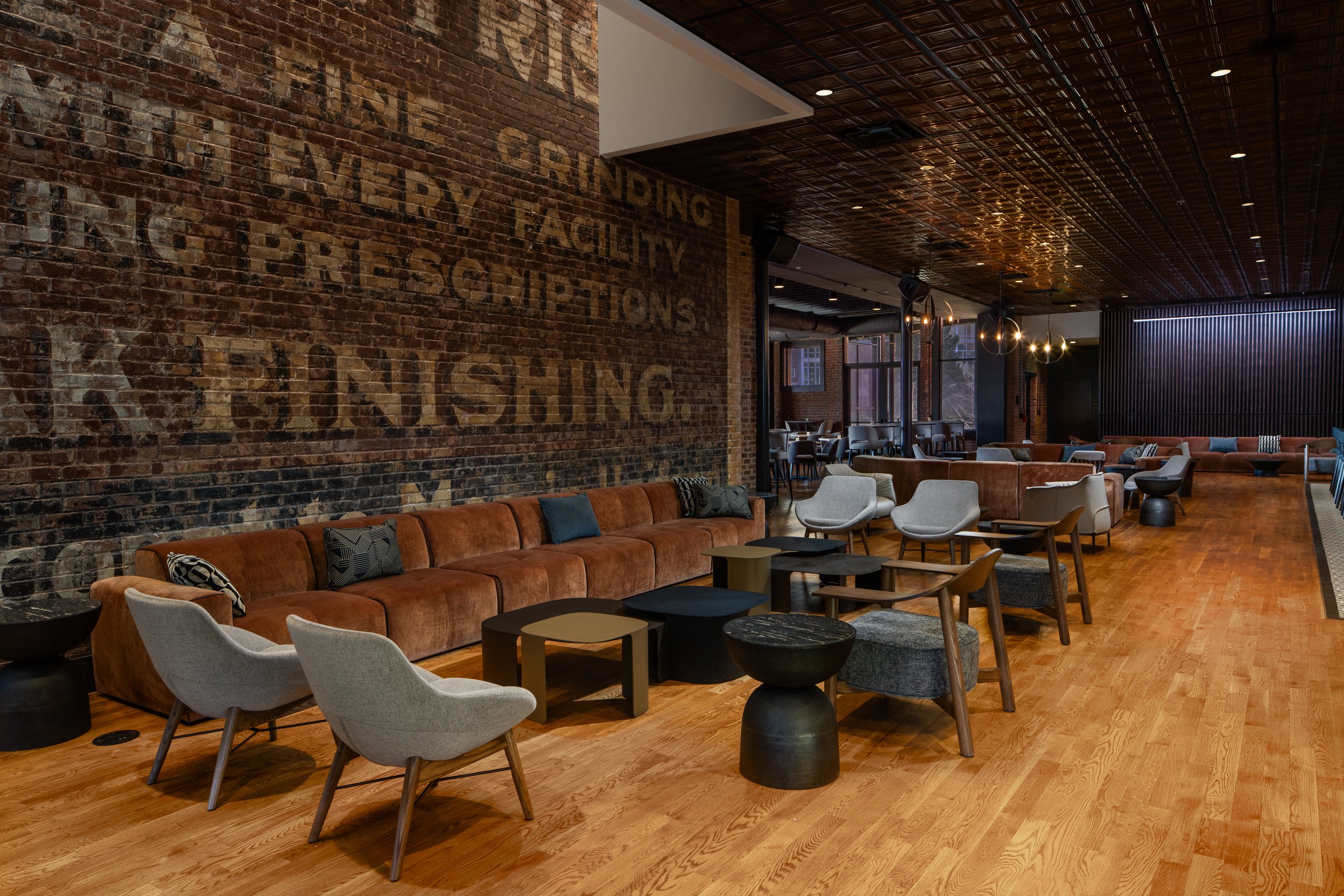 Long banquette and lounge seating beneath the building's original painted brick advertising signage, with a pressed tin ceiling and warm oak floors anchoring the room.