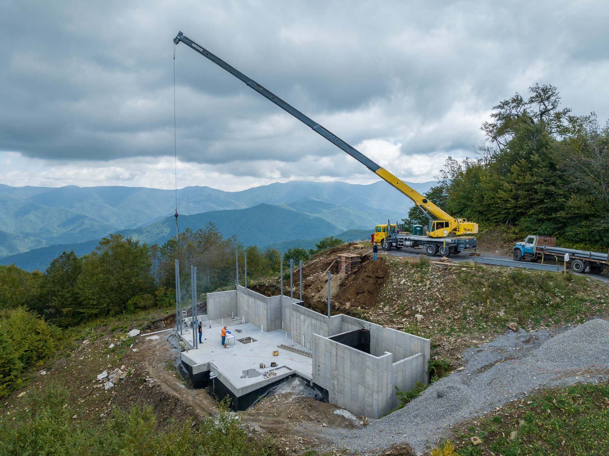 Aerial view of a mountain construction site with concrete foundation walls, workers, a yellow crane, gravel roads, and layered Blue Ridge mountain views.