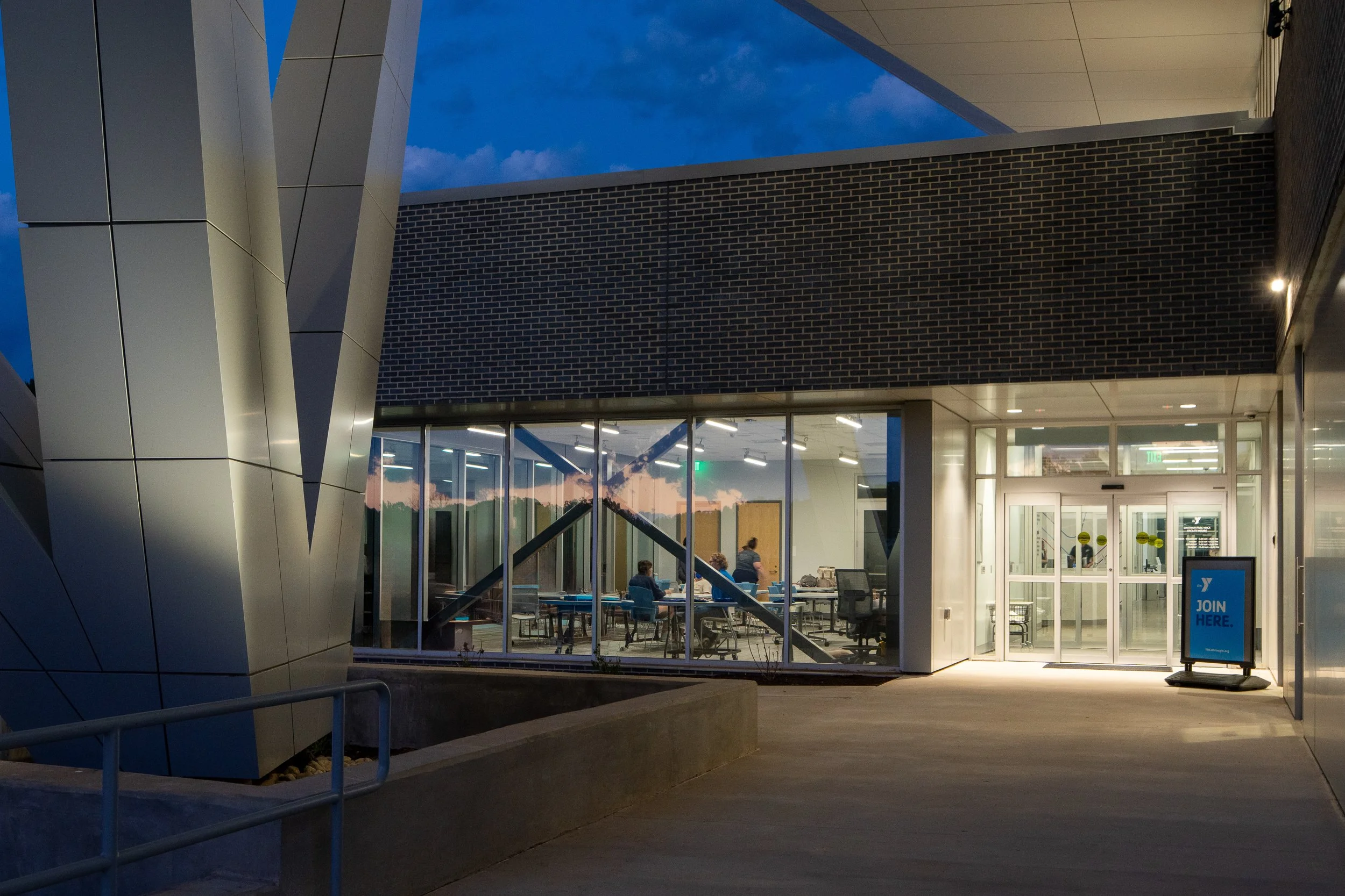 Glass-walled meeting rooms and bright entry doors create a clear evening approach to the YMCA lobby.