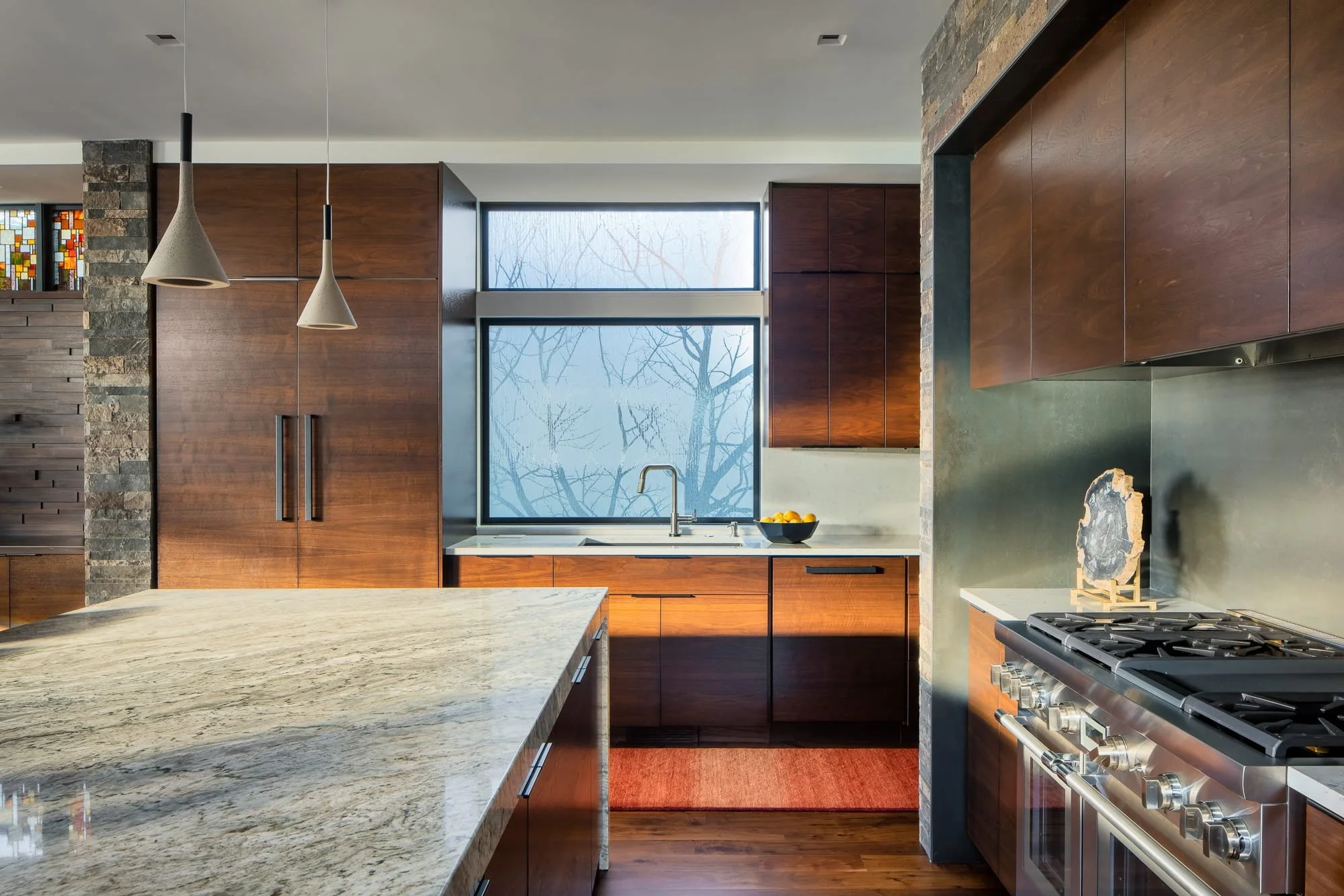Modern kitchen with rich wood cabinets, stone accent walls, a large marble island, pendant lights, and sunlight through frosted windows.