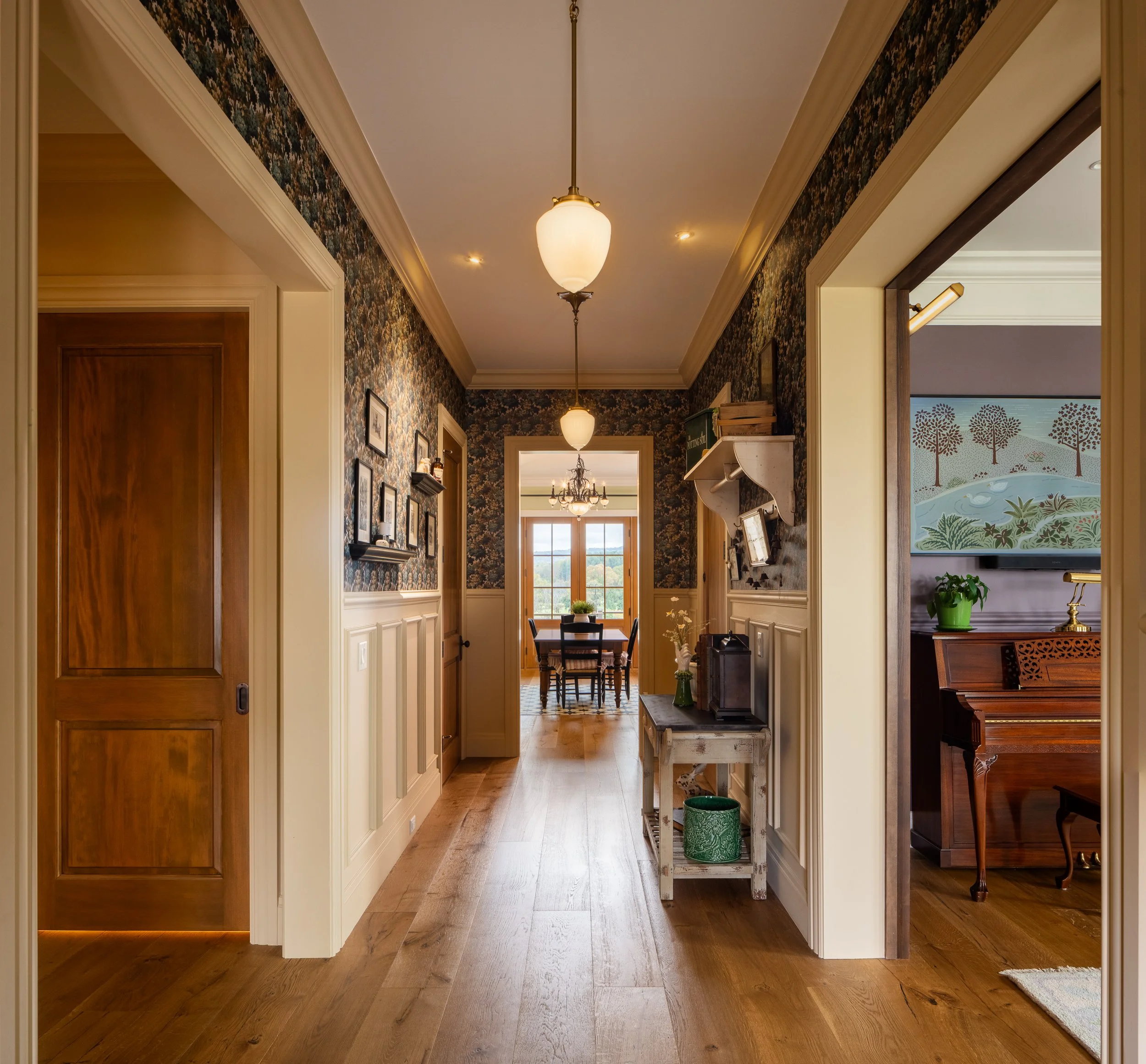 Long hallway with patterned wallpaper, paneled wainscoting, pendant lights, and warm wood floors