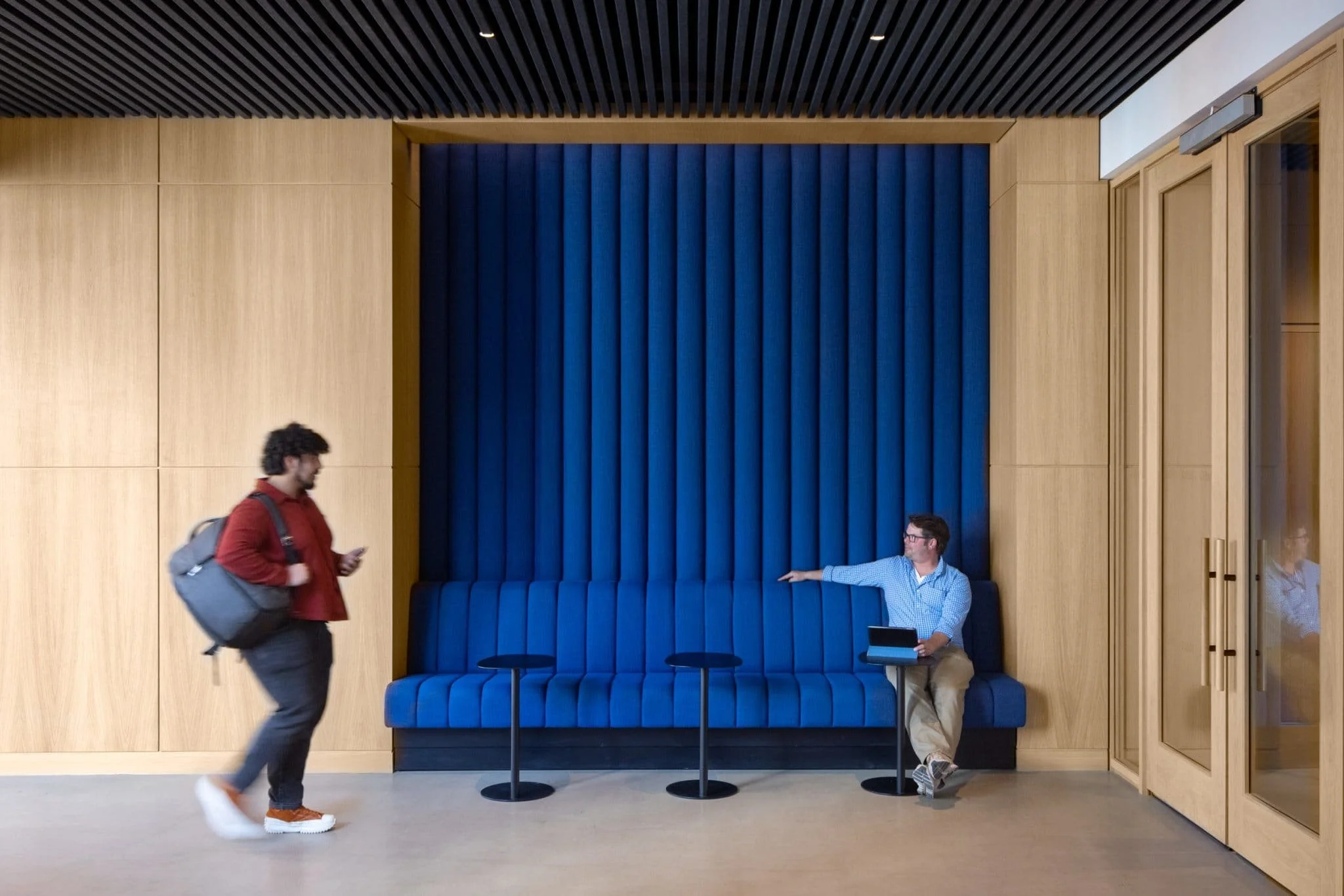 Contemporary lobby interior with a bright blue upholstered seating nook, wood wall panels, black slatted ceiling, and people using the space.