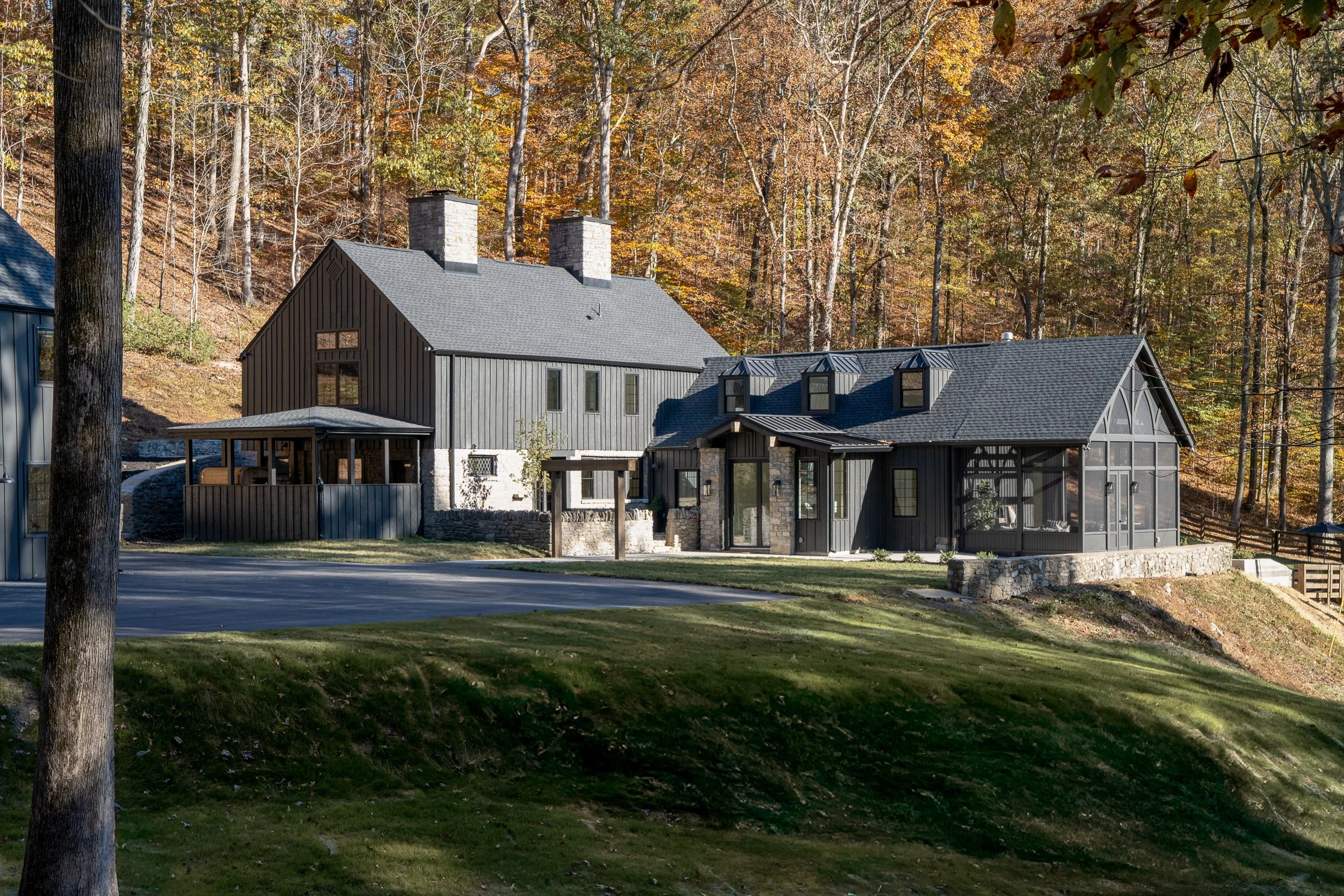 Exterior view of the renovated farmhouse with dark vertical siding, stone chimneys, screened porch, and wooded hillside.