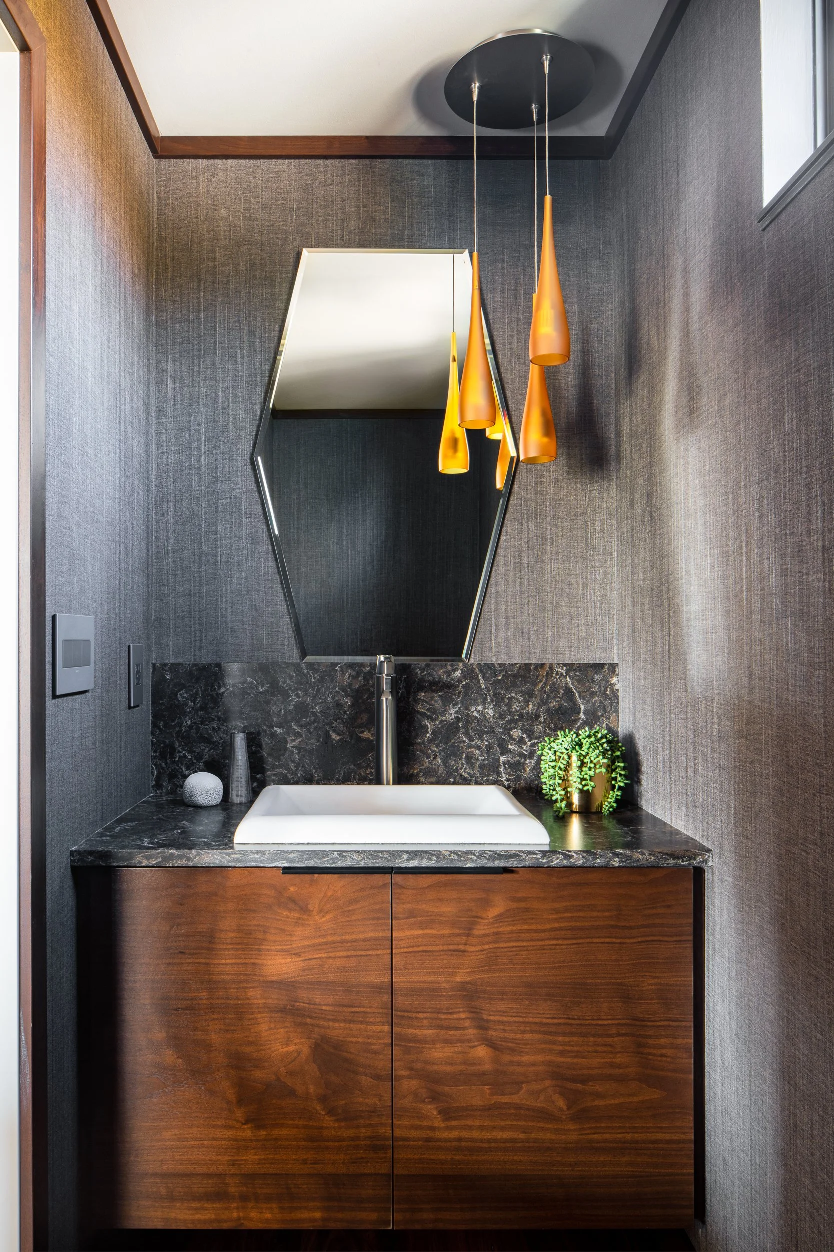 Powder room with wood vanity, dark stone countertop, gray wallcovering, and sculptural pendant lights.