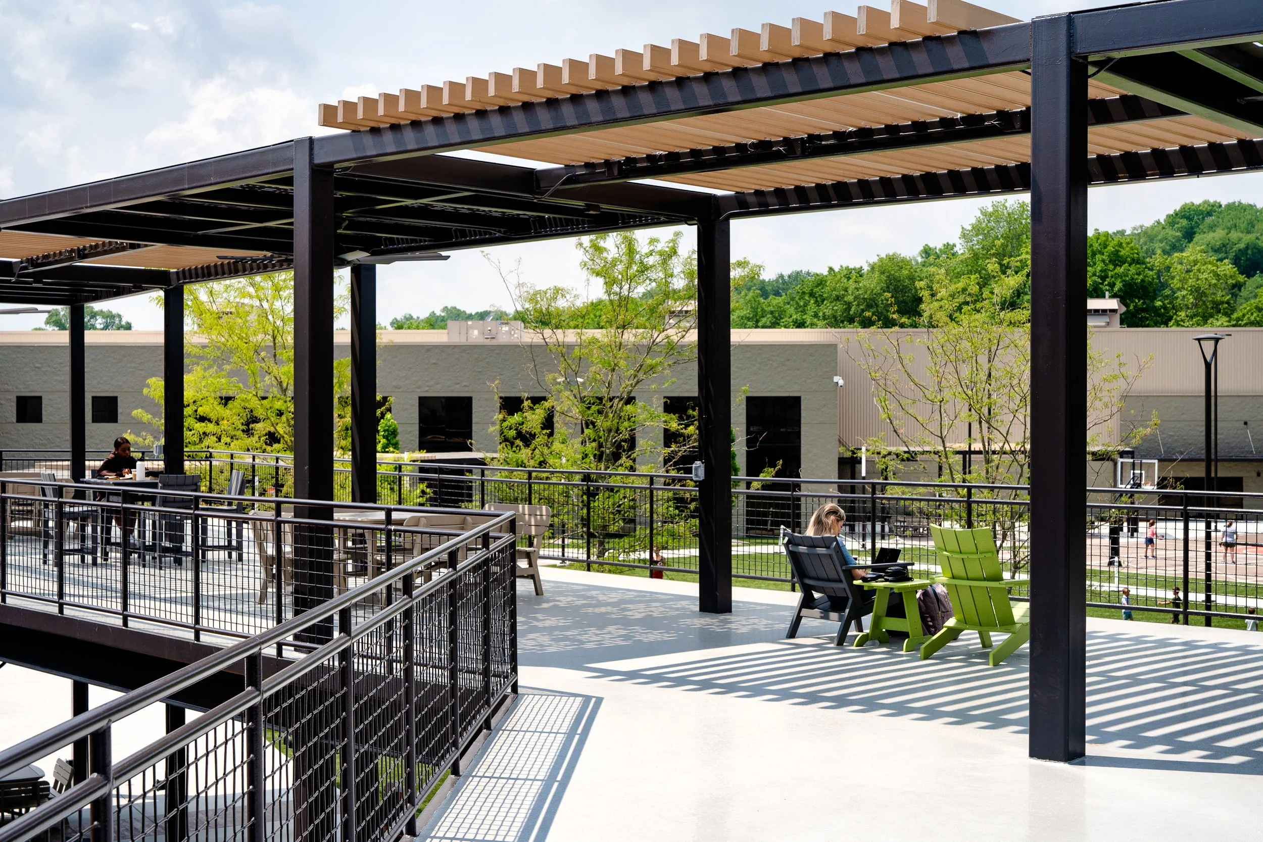 Outdoor terrace with black pergola, seating areas, railings, and landscaped commercial buildings beyond.