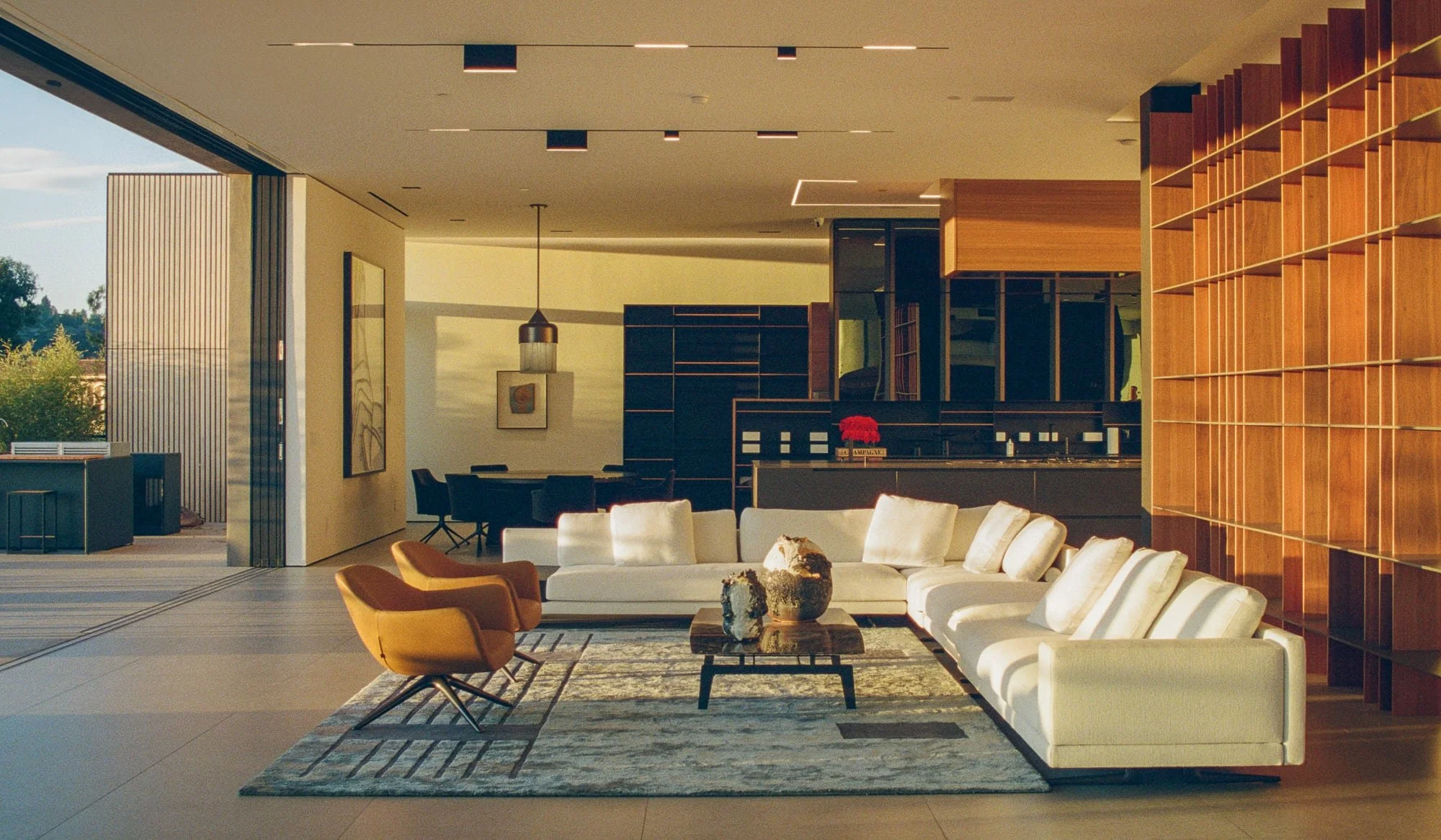 Modern living room with beige sofa, yellow armchair, and coffee table on a rug, open to a dining area with dark chairs and art on the wall, large windows with outdoor view.
