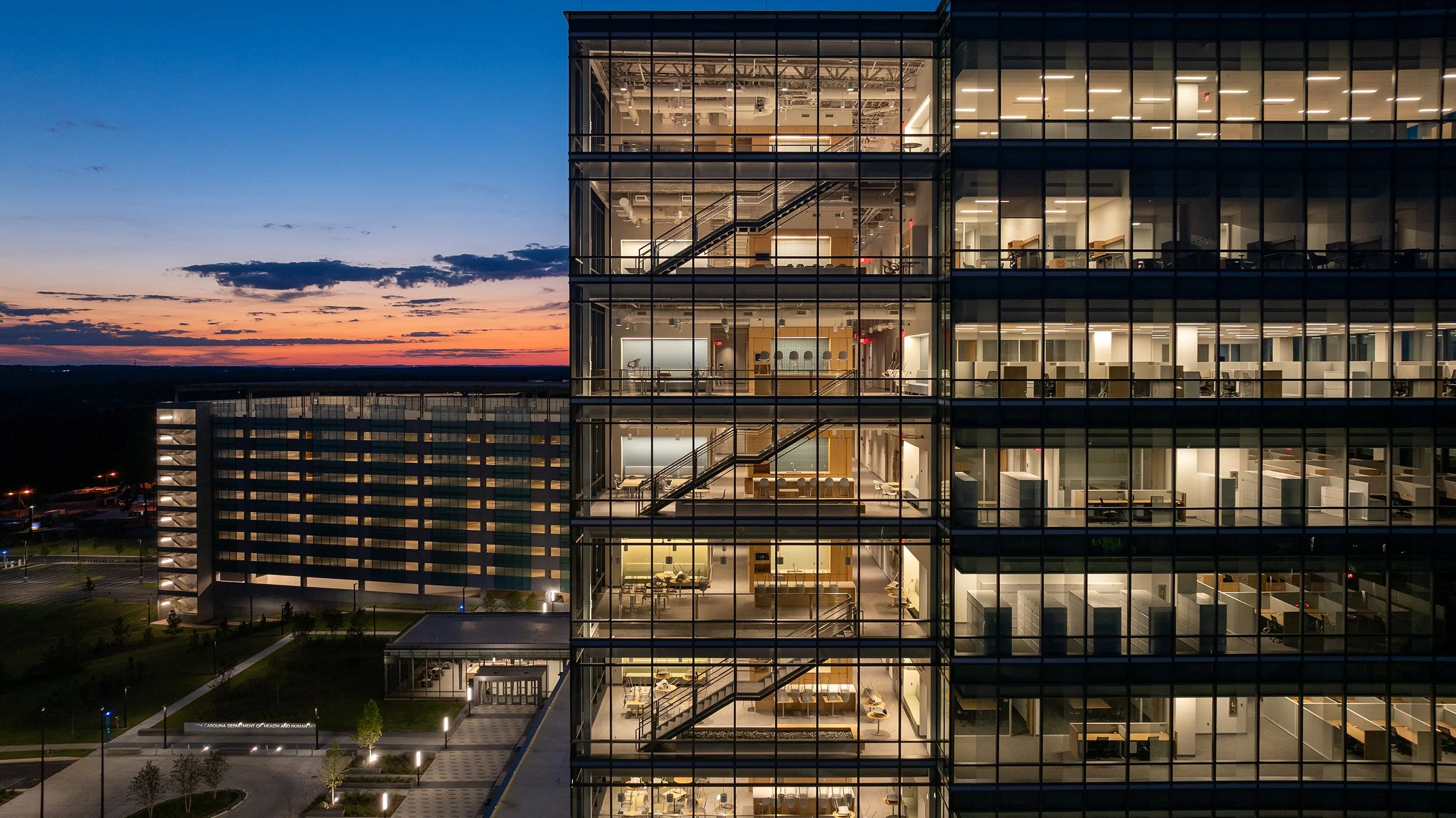 Close aerial view of the glass curtain wall at dusk, revealing the building's stacked open stairs, shared landings, and lit interiors at every level.