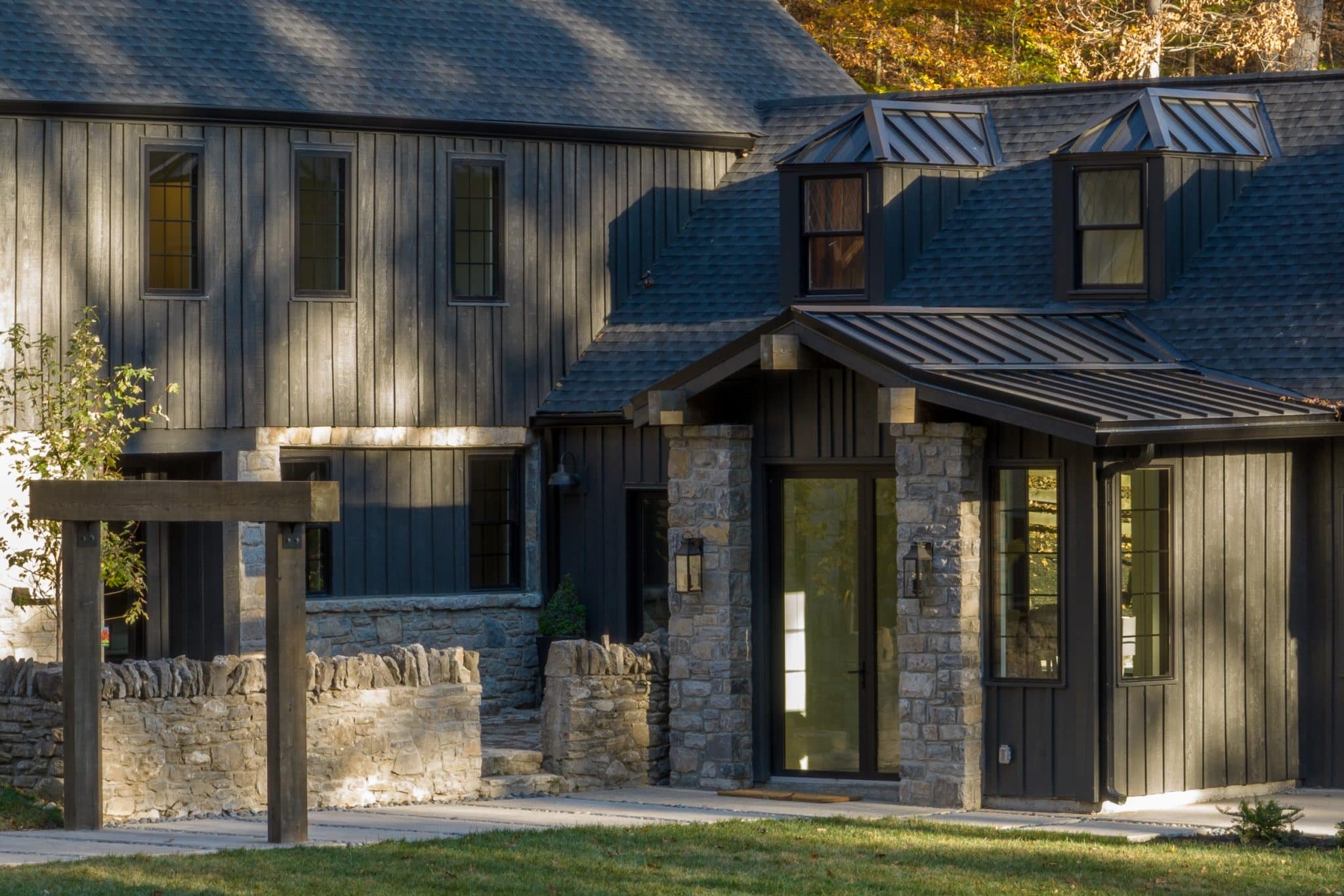 Exterior view of a modern house with stone and wood siding, large windows, and a metal roof, surrounded by trees with autumn foliage.