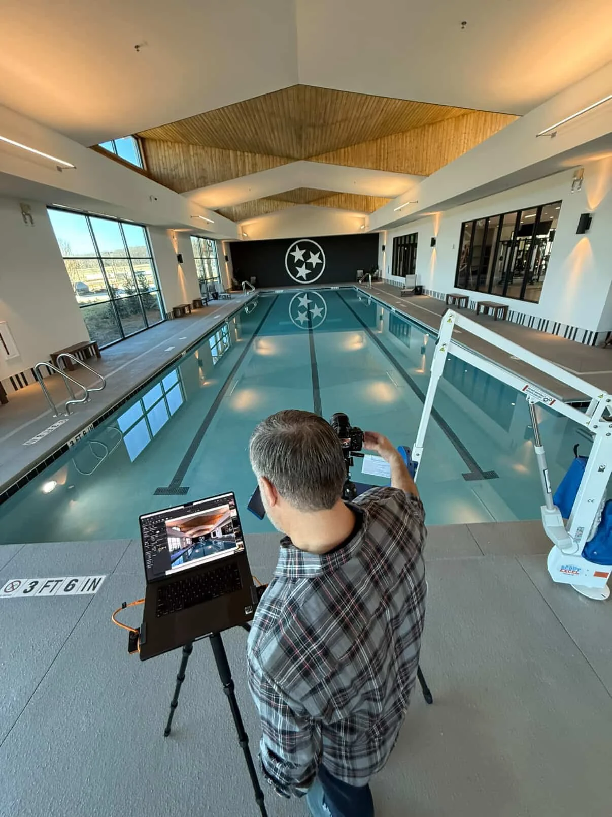 Inside an indoor swimming pool with a man taking photographs, no lifeguard or swimmers present, a logo with three stars inside a circle on the back wall, windows on the left side, and pool safety signs on the floor.