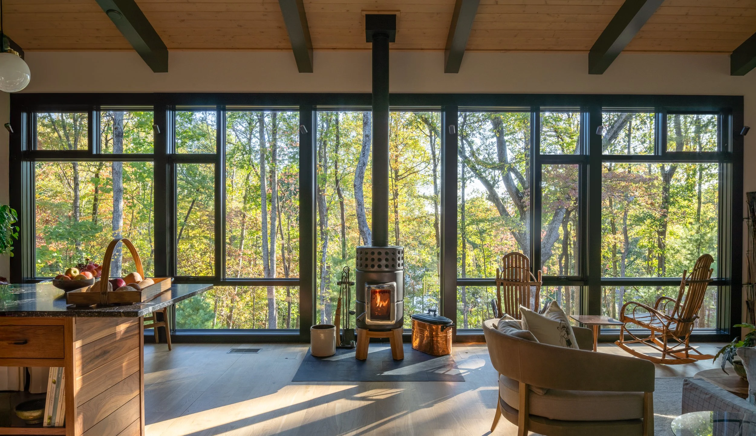Round wood stove centered in front of a tall wall of windows overlooking the trees.