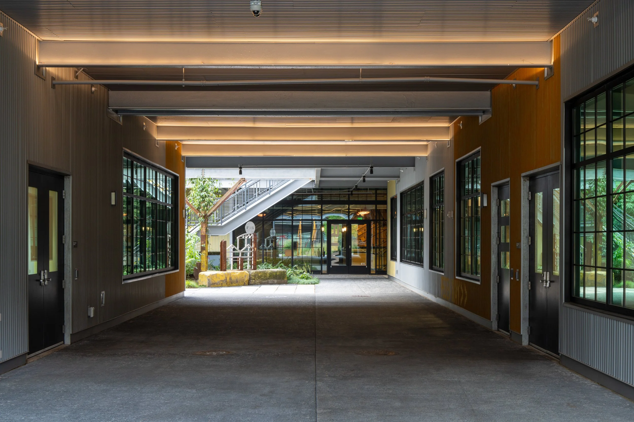 Covered passageway under the building with corrugated metal walls, polished concrete floor, and a view through to the lit North Lobby with its preserved rusted steel column beyond.