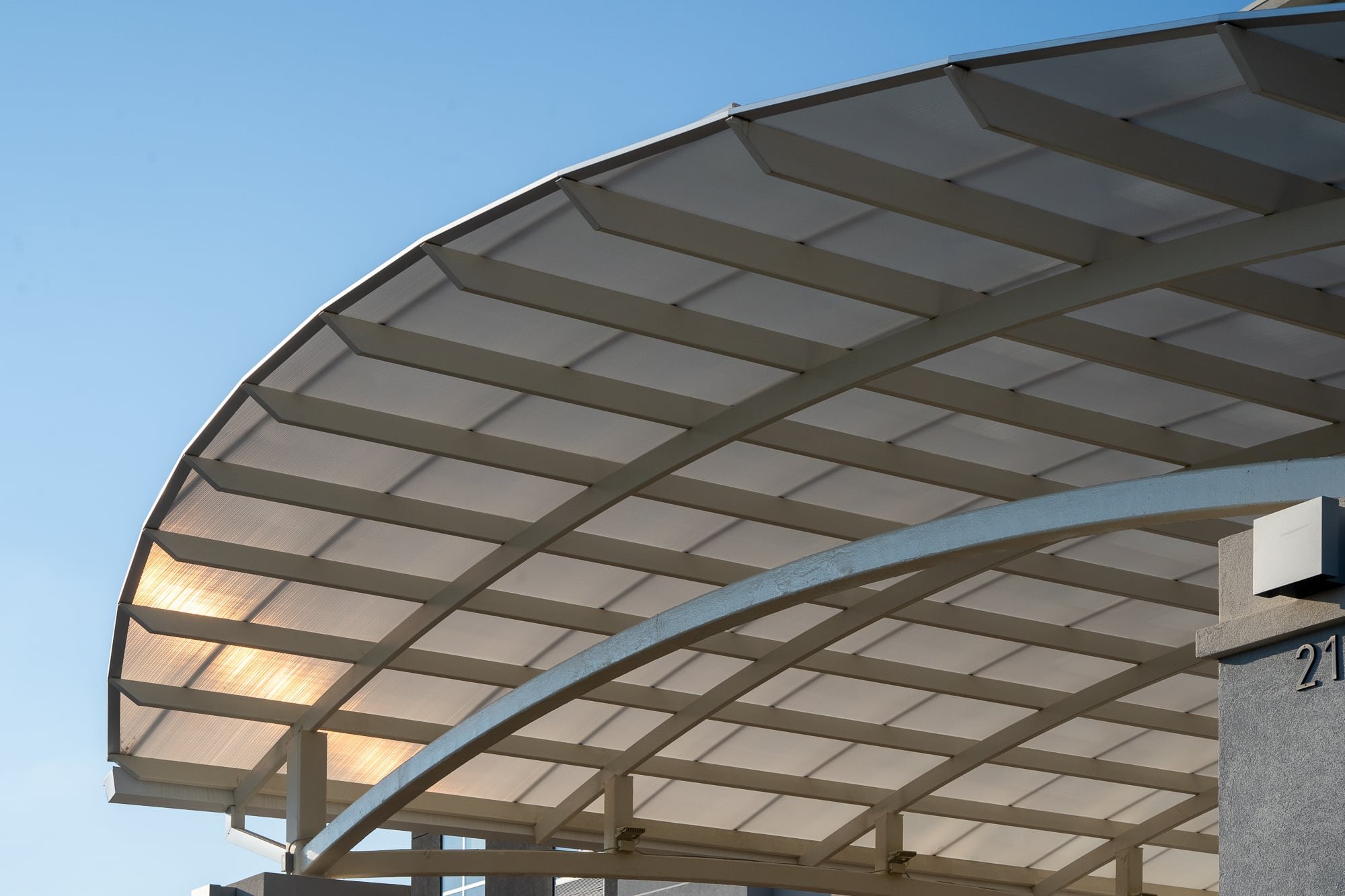 Close view of a curved architectural canopy with framing and translucent polycarbonate panels against a clear blue sky.