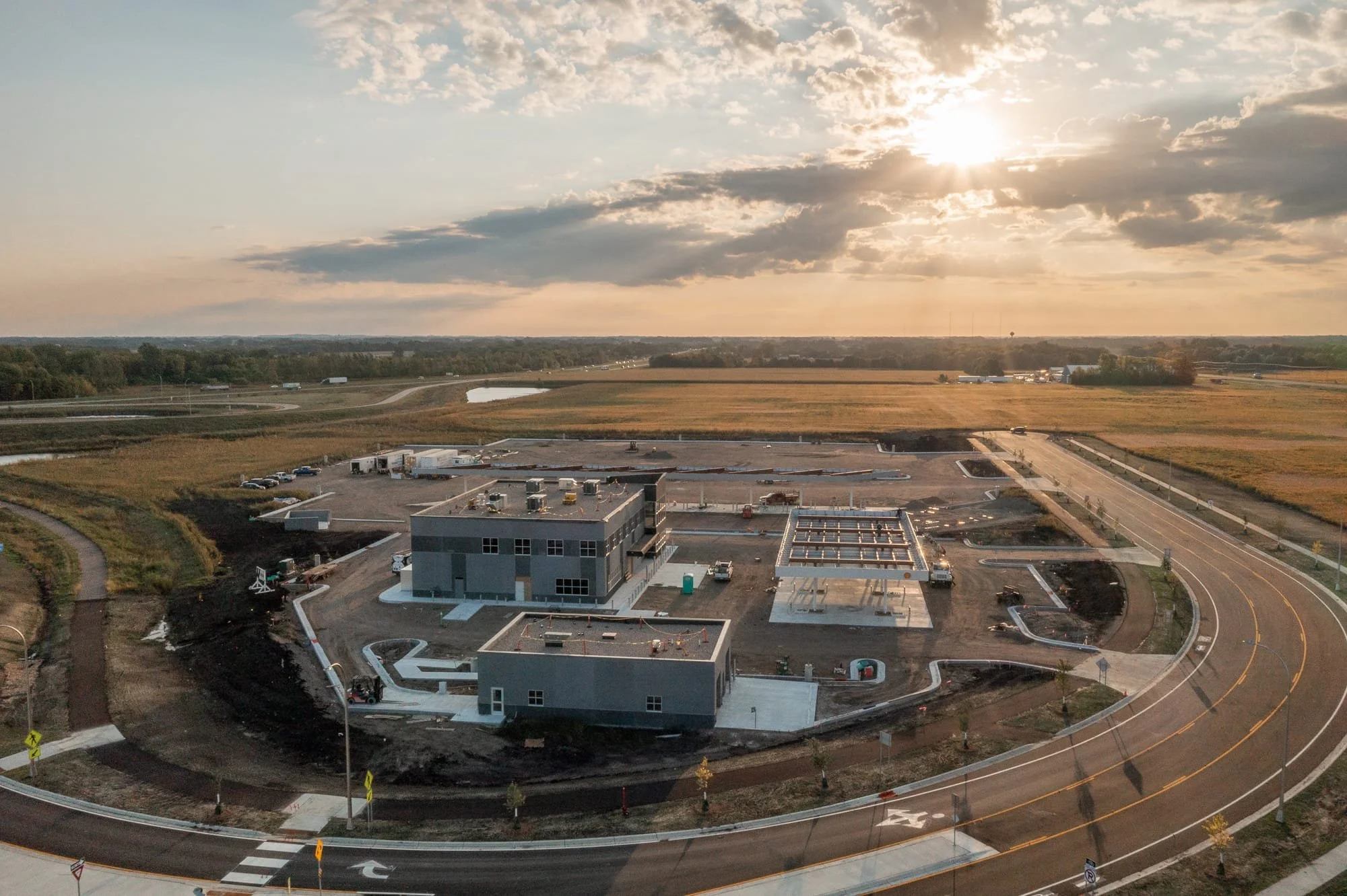 Drone view of a commercial construction site at sunset, showing partially built structures, roads, fields, and long evening shadows.