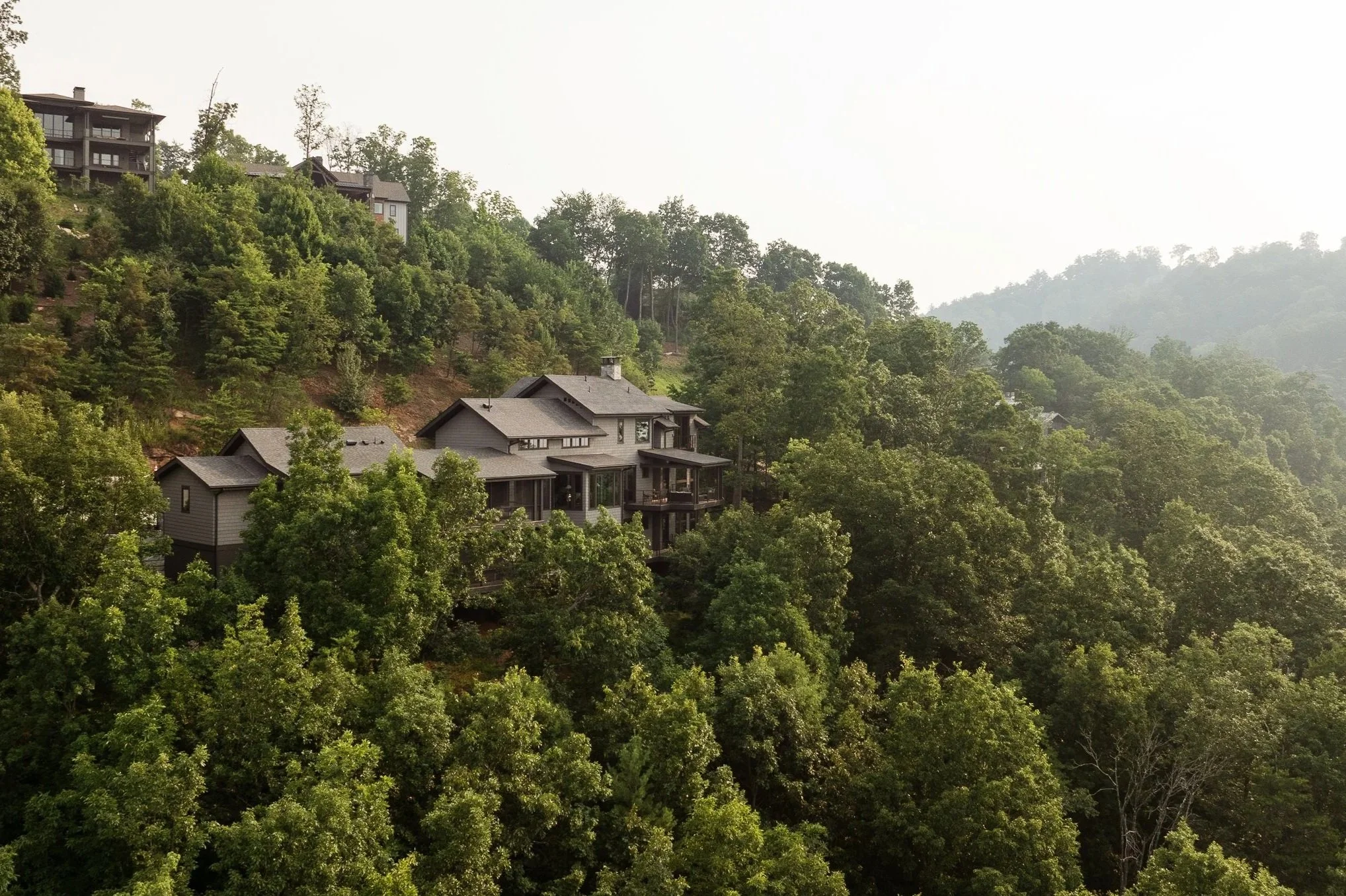 Houses built on a hillside surrounded by dense green trees with a hazy sky in the background.