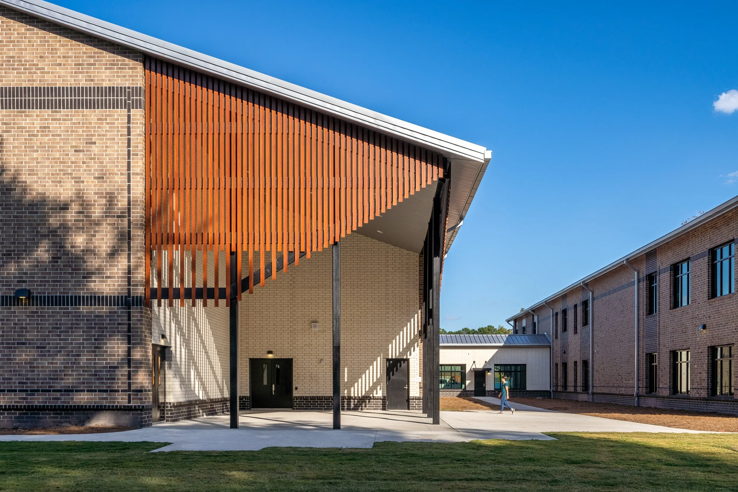 Covered exterior walkway with vertical wood screening, brick walls, metal roof, and a person walking between school buildings.