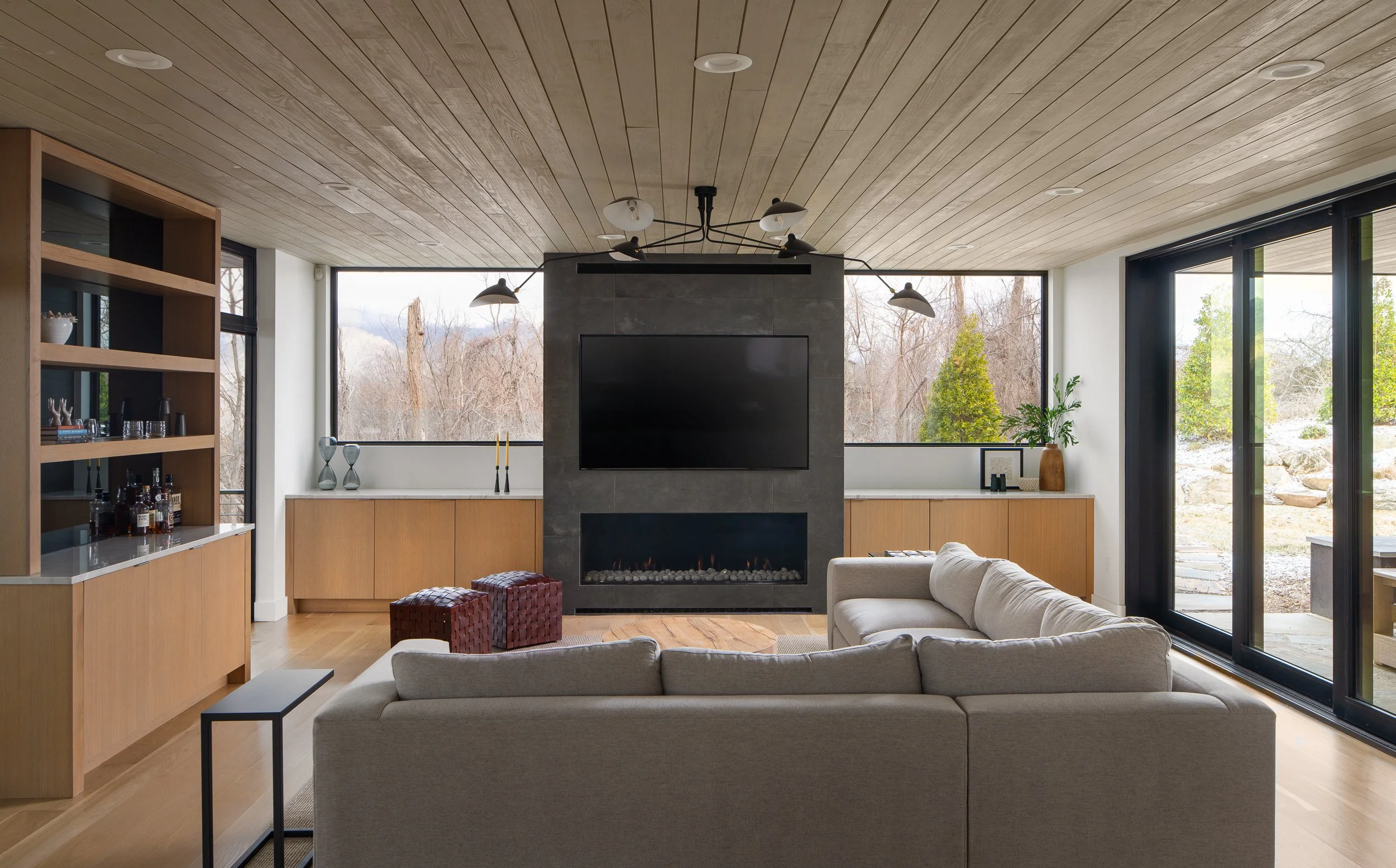 Living room with a wood ceiling, central fireplace wall, built-in cabinetry, and large windows facing the trees.