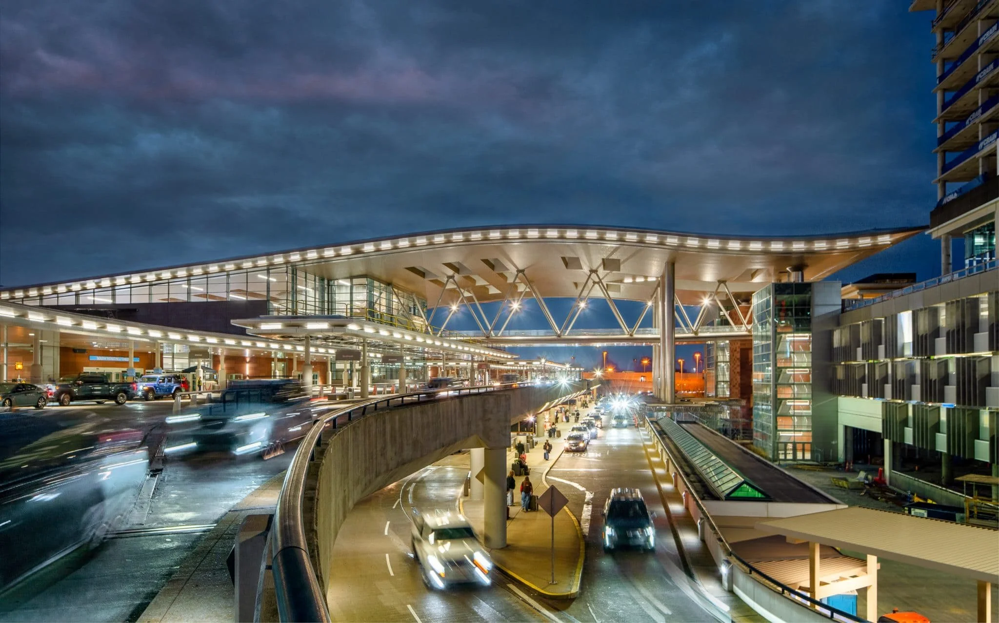Night architectural photo of an airport terminal roadway with moving traffic, elevated lanes, glass stair towers, and a broad illuminated canopy.