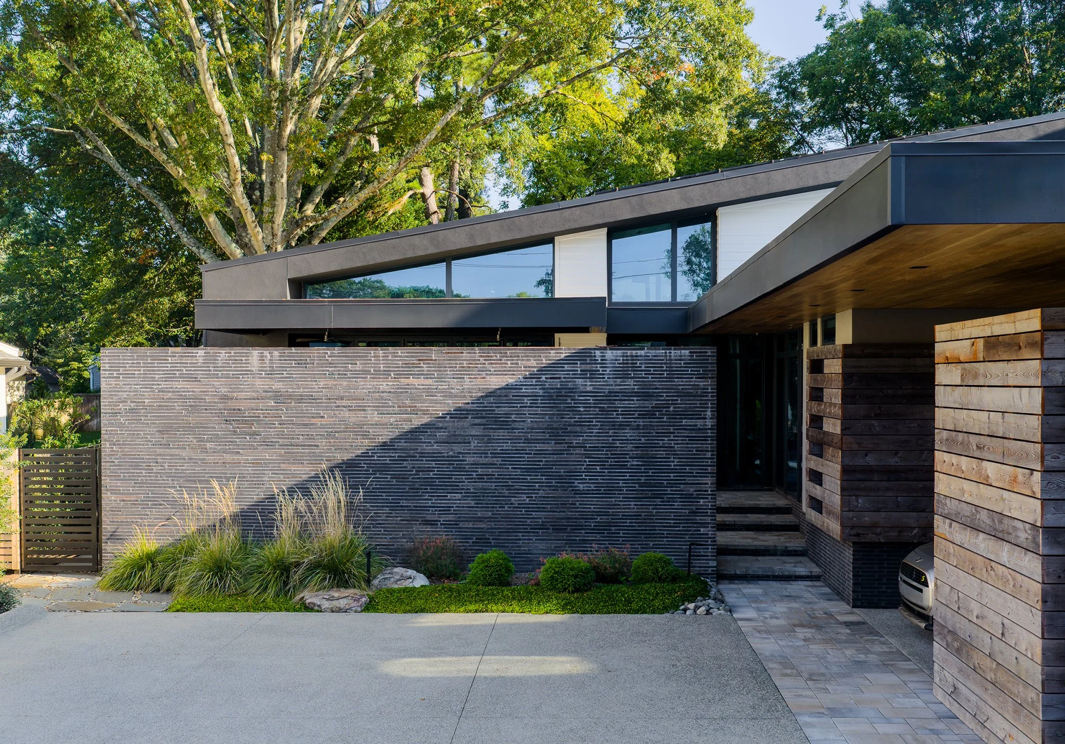 A sloping roof with high windows extends above a dark brick wall and recessed entrance beside horizontal wood siding.