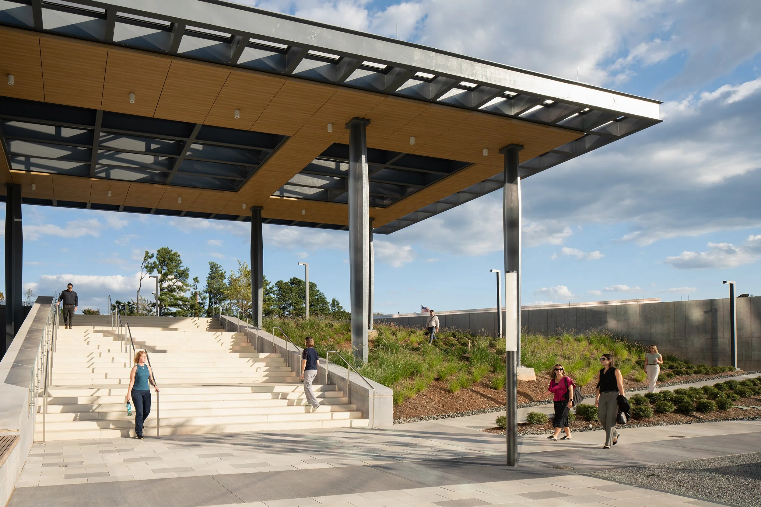 Wood-lined entry canopy supported by slim steel columns, with broad concrete steps descending to the plaza and visitors making their way to and from the building.