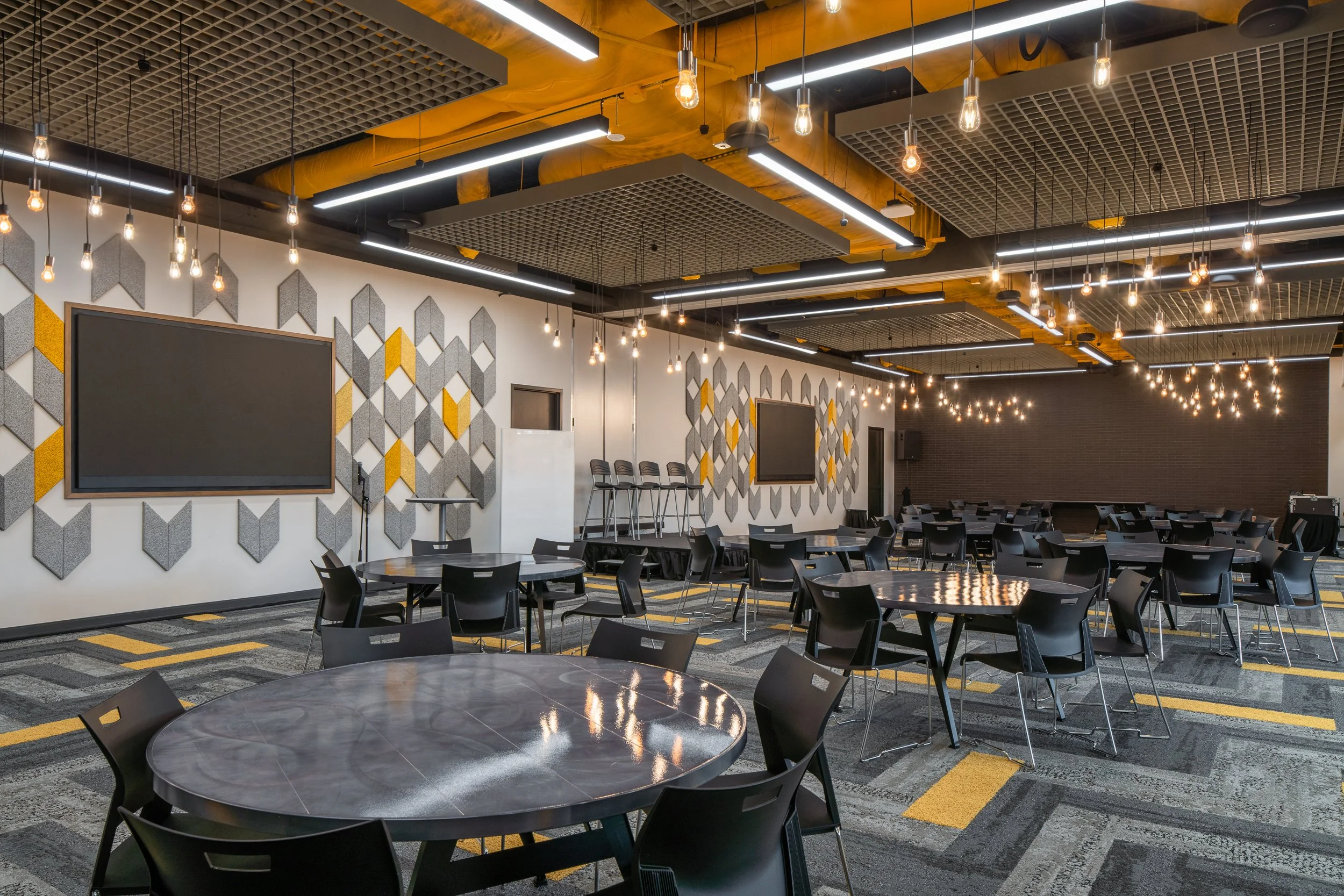 Spacious event hall with geometric wall panels, black chairs, and warm pendant lighting.