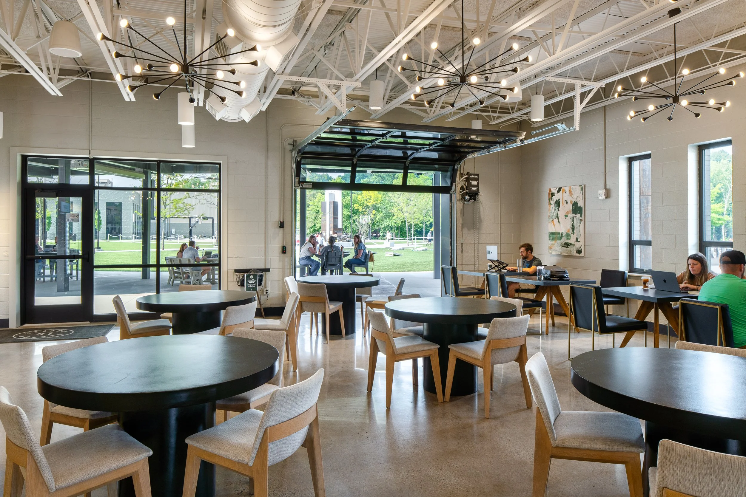 Spacious cafe interior with modern black tables, wooden chairs, and open garage door leading to outdoor seating.