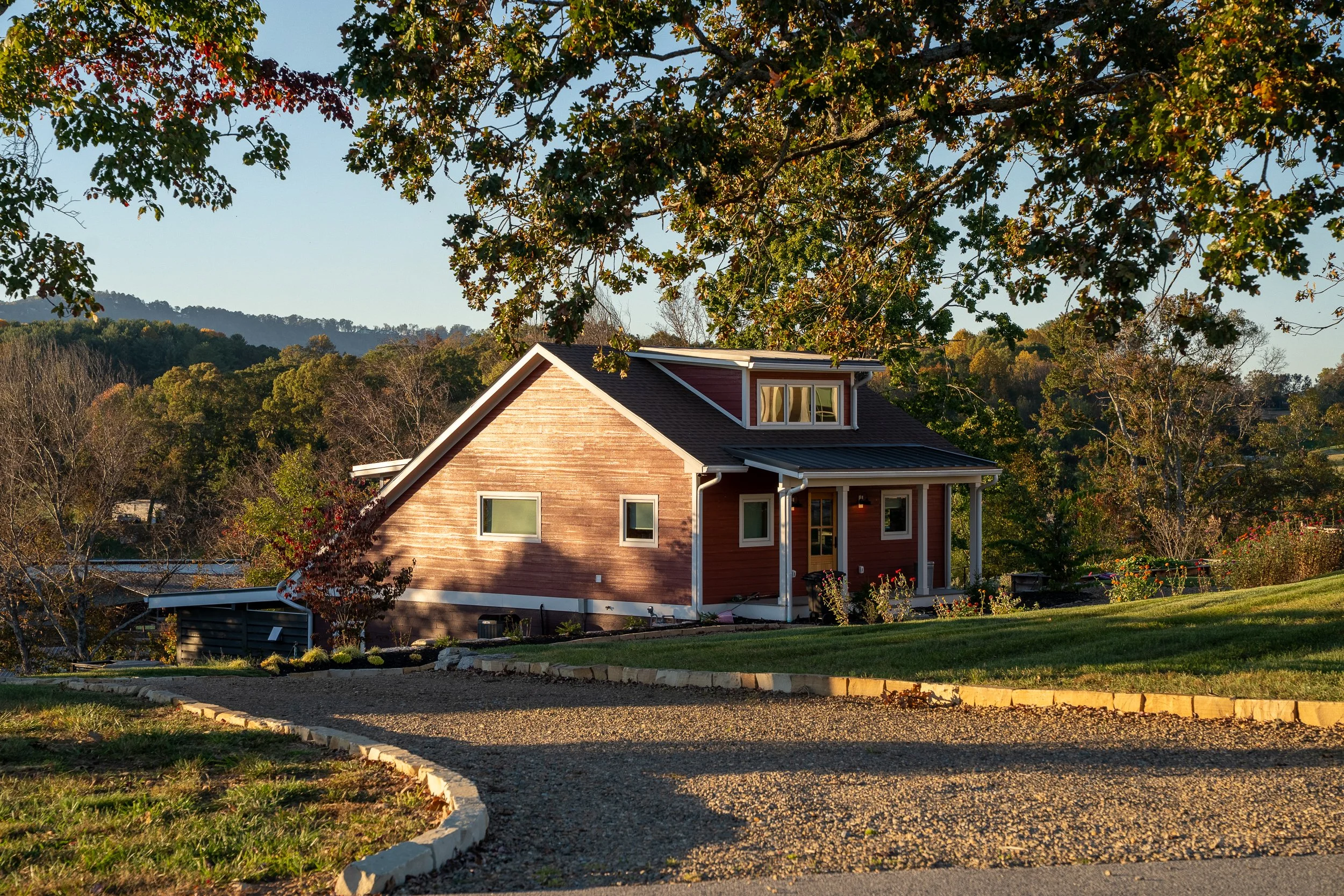 A red wooden house with white trim, large windows, and a small balcony, surrounded by trees and a neatly landscaped yard with a curved gravel driveway.