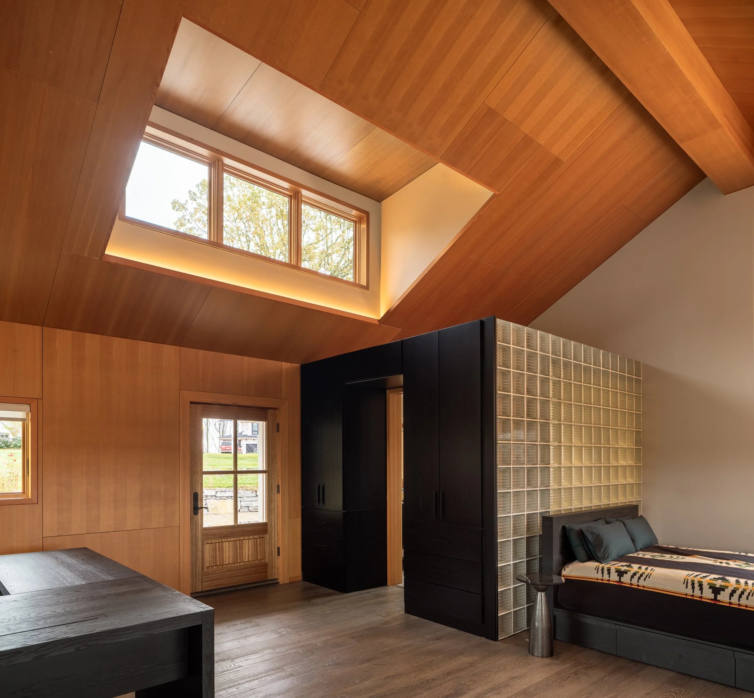 Modern loft bedroom with glass block wall, black cabinetry, low bed, and wood-paneled ceiling.
