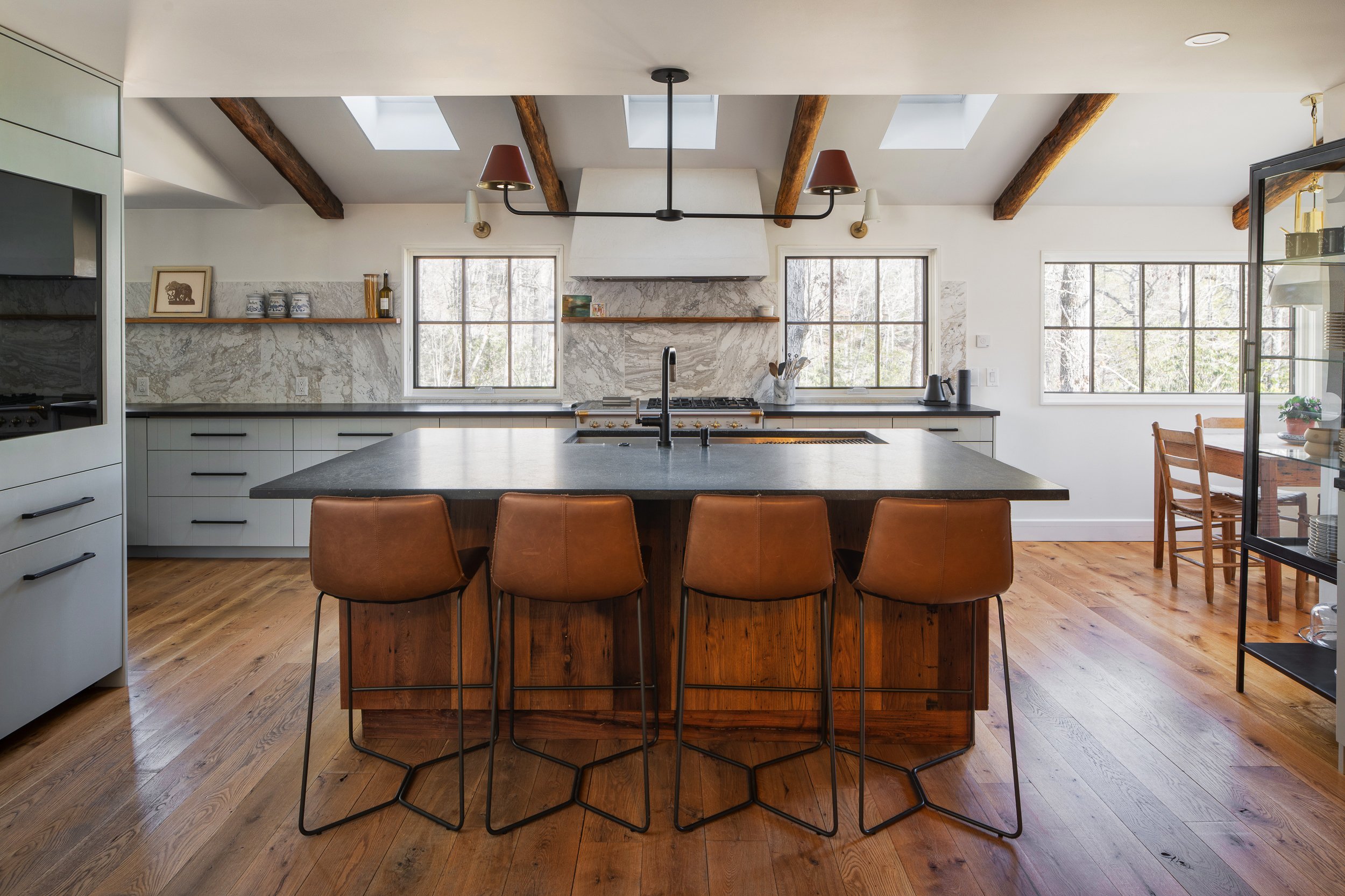 Bright kitchen with a large dark island, leather stools, marble backsplash, skylights, and exposed wood beams.