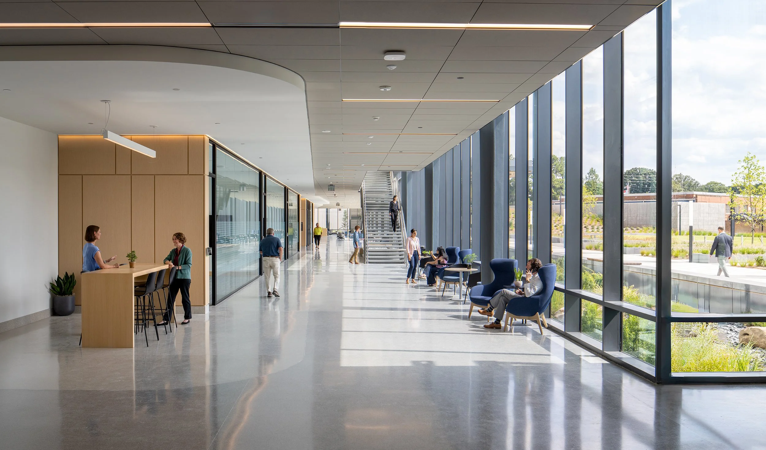 Wide concourse with polished concrete floors, lounge seating clustered along the full-height curtain wall, a curved wood-clad service point on the left, and an open stair in the distance.